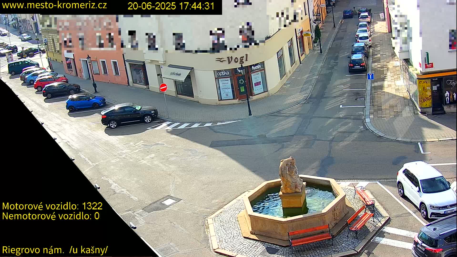 A sunny town square with parked cars, a stone fountain with a statue, and several benches.