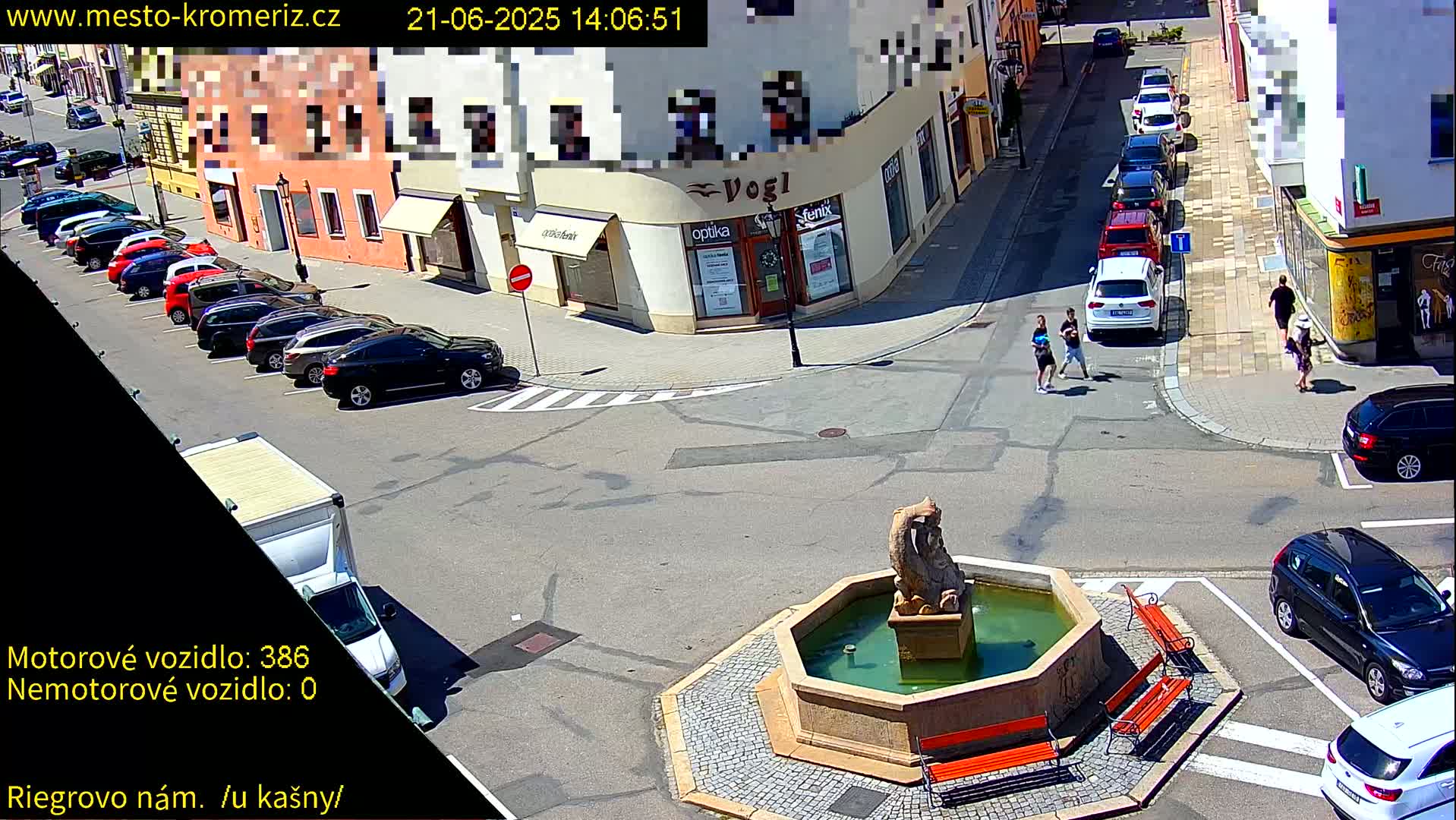 A sunny town square with a fountain, several parked cars, and a few pedestrians is viewed from above.