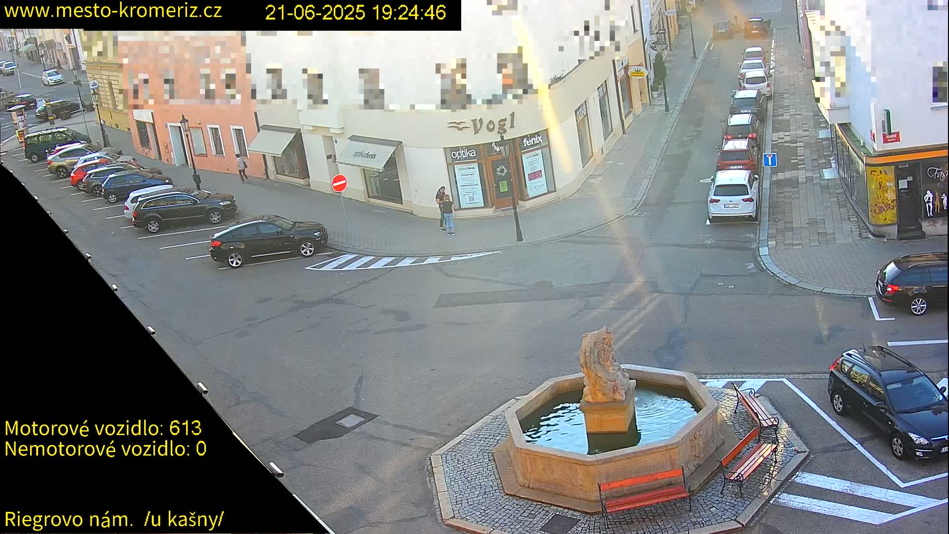 A sunny town square scene shows several parked cars surrounding a stone fountain with benches.