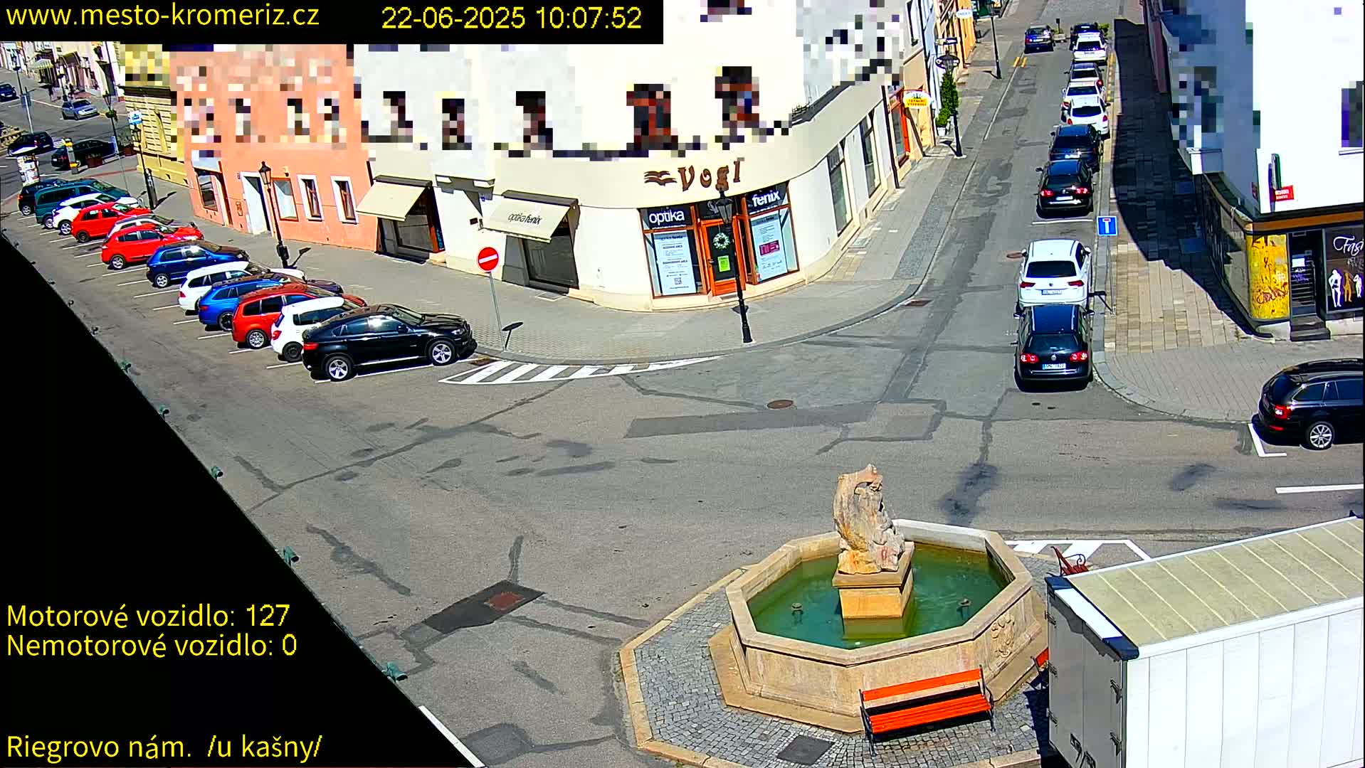 A street scene on a sunny day shows several parked cars lining a street, a fountain in a town square, and several more cars driving on the intersecting street.
