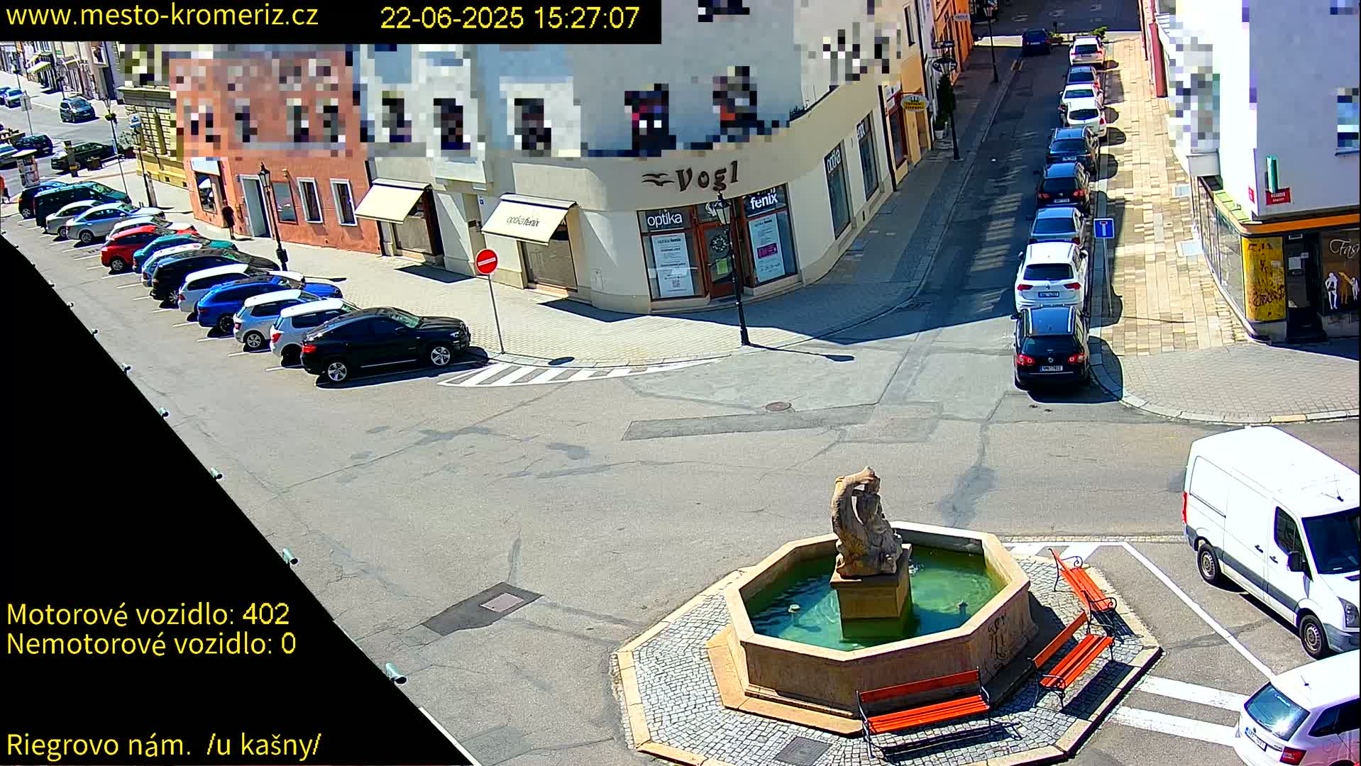 A sunny town square with a fountain, several parked cars, and a few vehicles on the road.