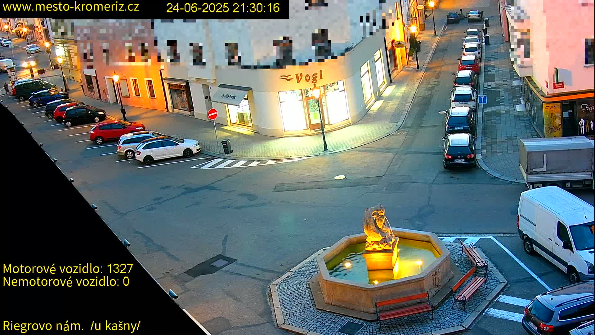 A town square at night, illuminated by streetlights, features a fountain with a statue, parked cars lining the streets, and a few cars driving by.