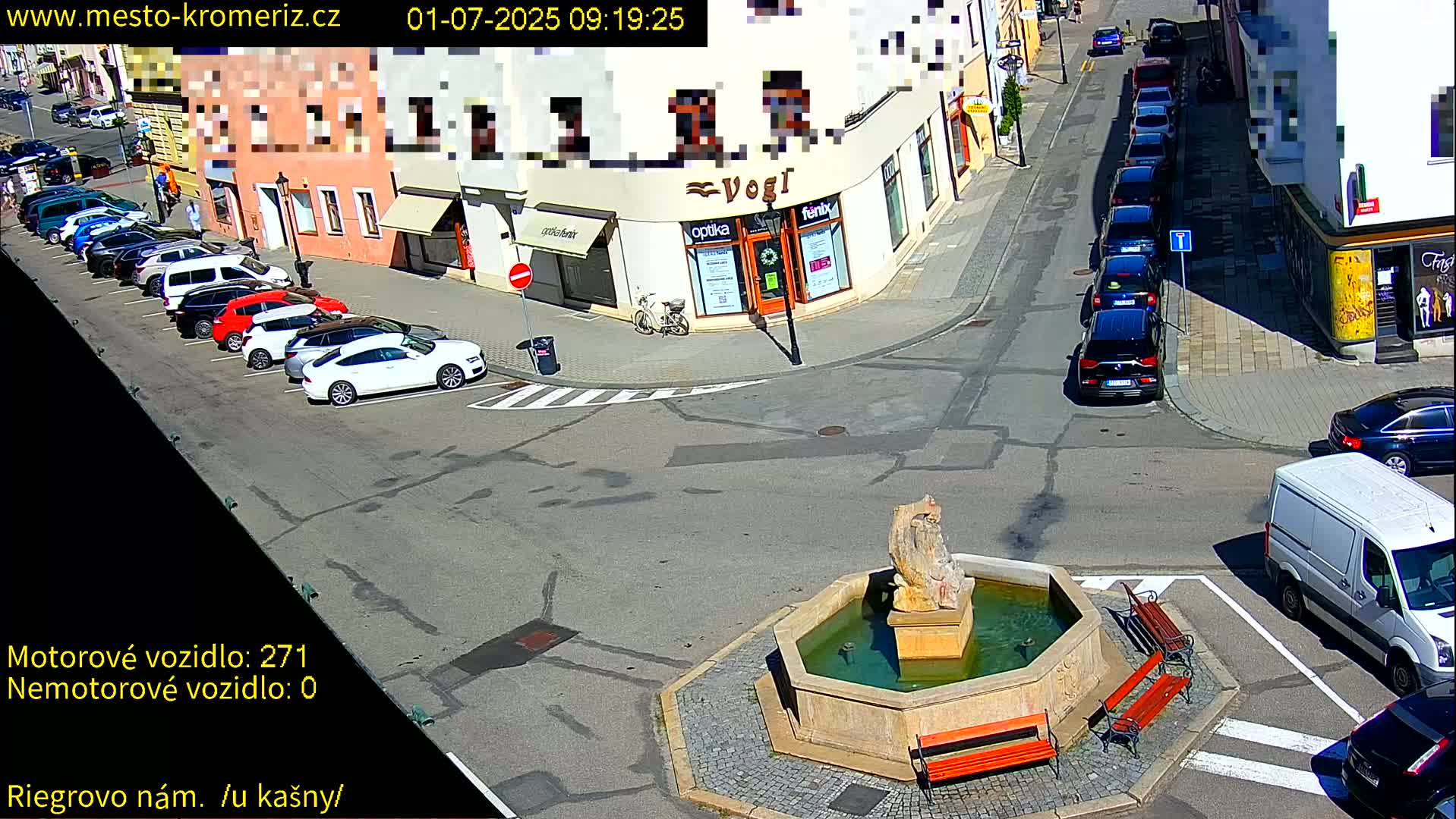 A sunny town square with a stone fountain at its center, surrounded by parked cars and buildings.