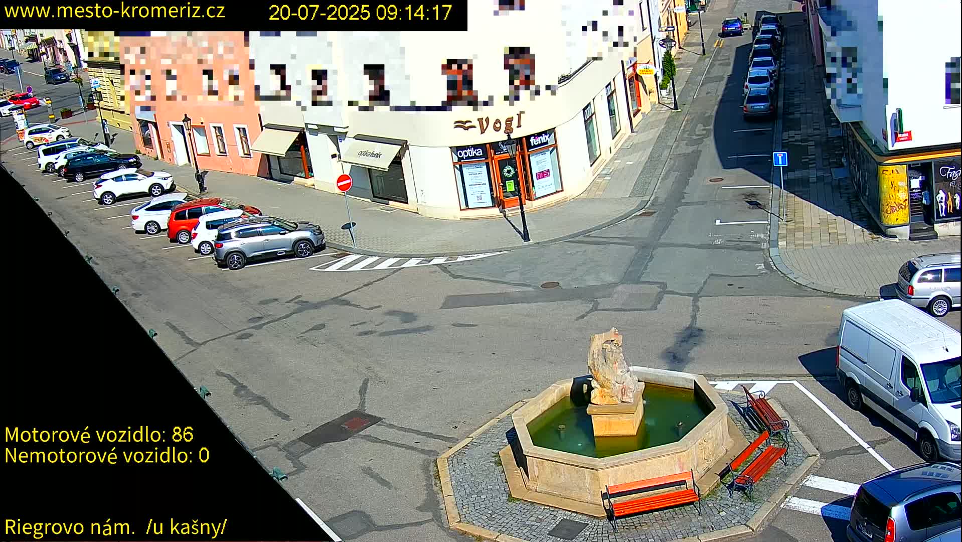 A sunny town square shows several parked cars surrounding a stone fountain with benches, and a white van is parked nearby.