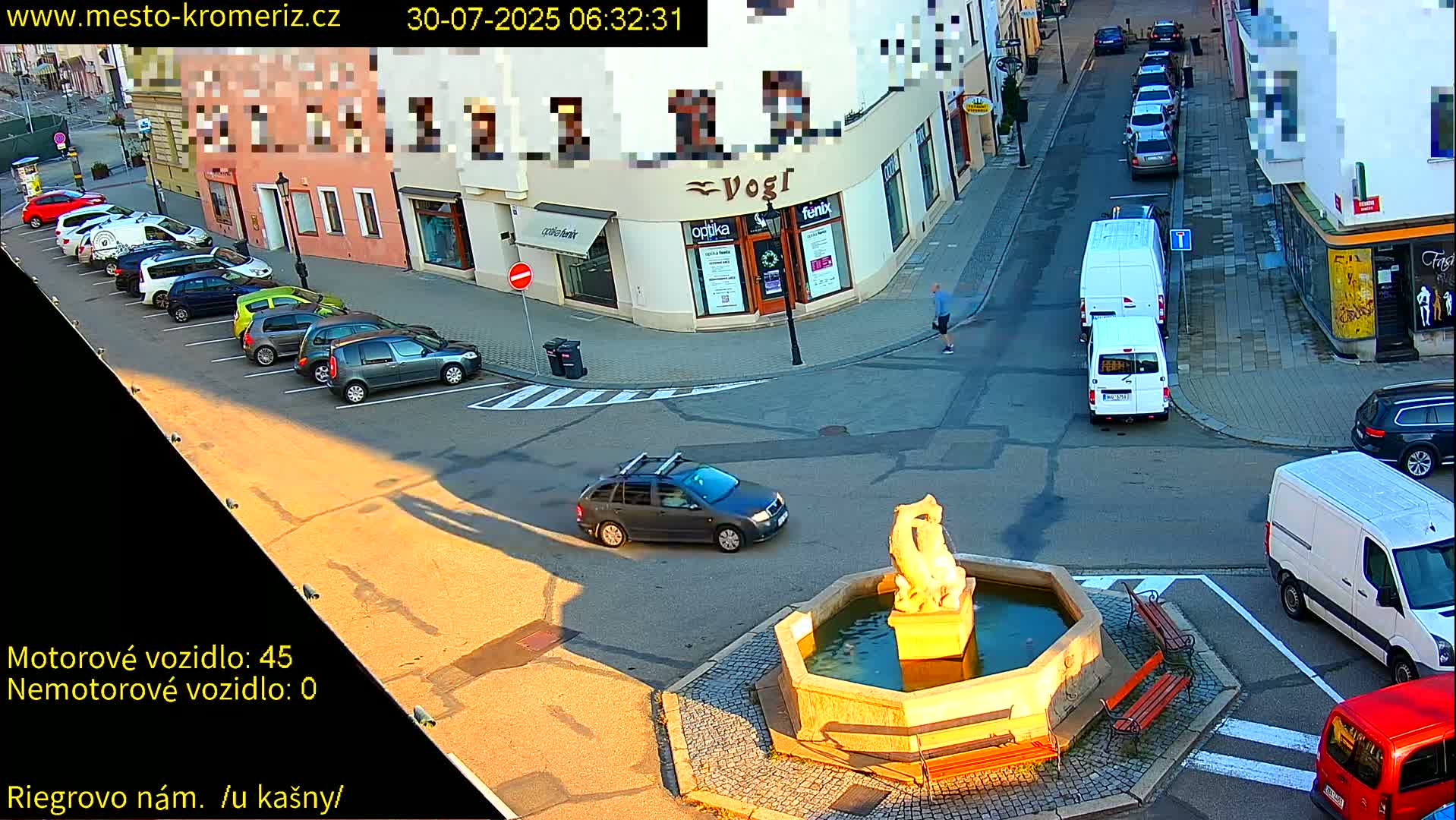A sunny town square shows several parked cars surrounding a stone fountain with benches, and a white van is parked nearby.