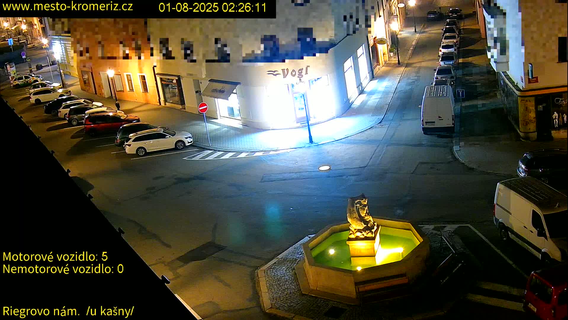 A nighttime street scene shows several parked cars around a fountain with a statue in the center, under well-lit conditions.