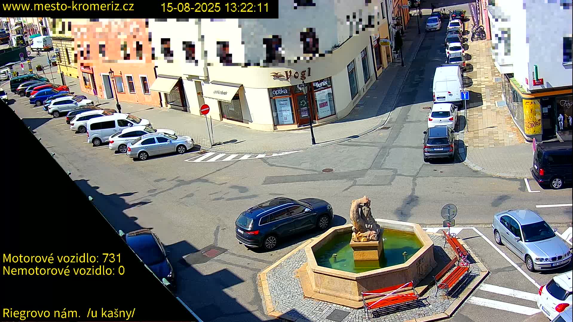 A sunny town square scene shows a fountain with people sitting nearby, cars parked along the streets, and buildings lining the square.