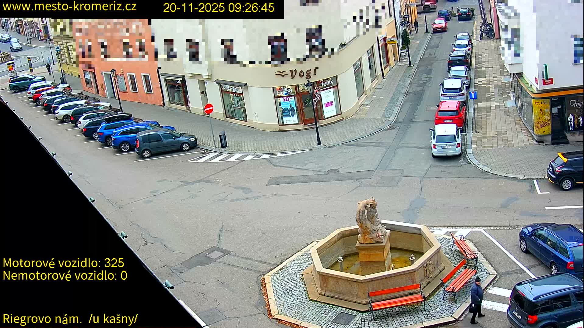 An overhead daytime view shows an urban intersection with a central stone fountain, surrounded by several parked vehicles, a few pedestrians, and multi-story buildings, all under an overcast sky.