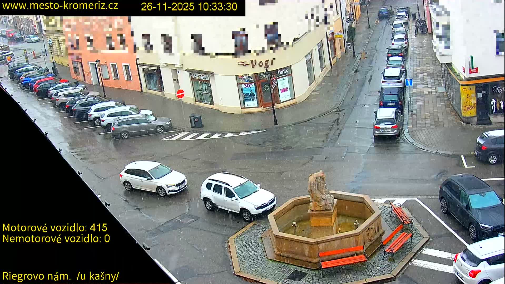 A rainy daytime street scene shows numerous cars parked and driving on wet roads, with a stone fountain and statue in the foreground, flanked by buildings.