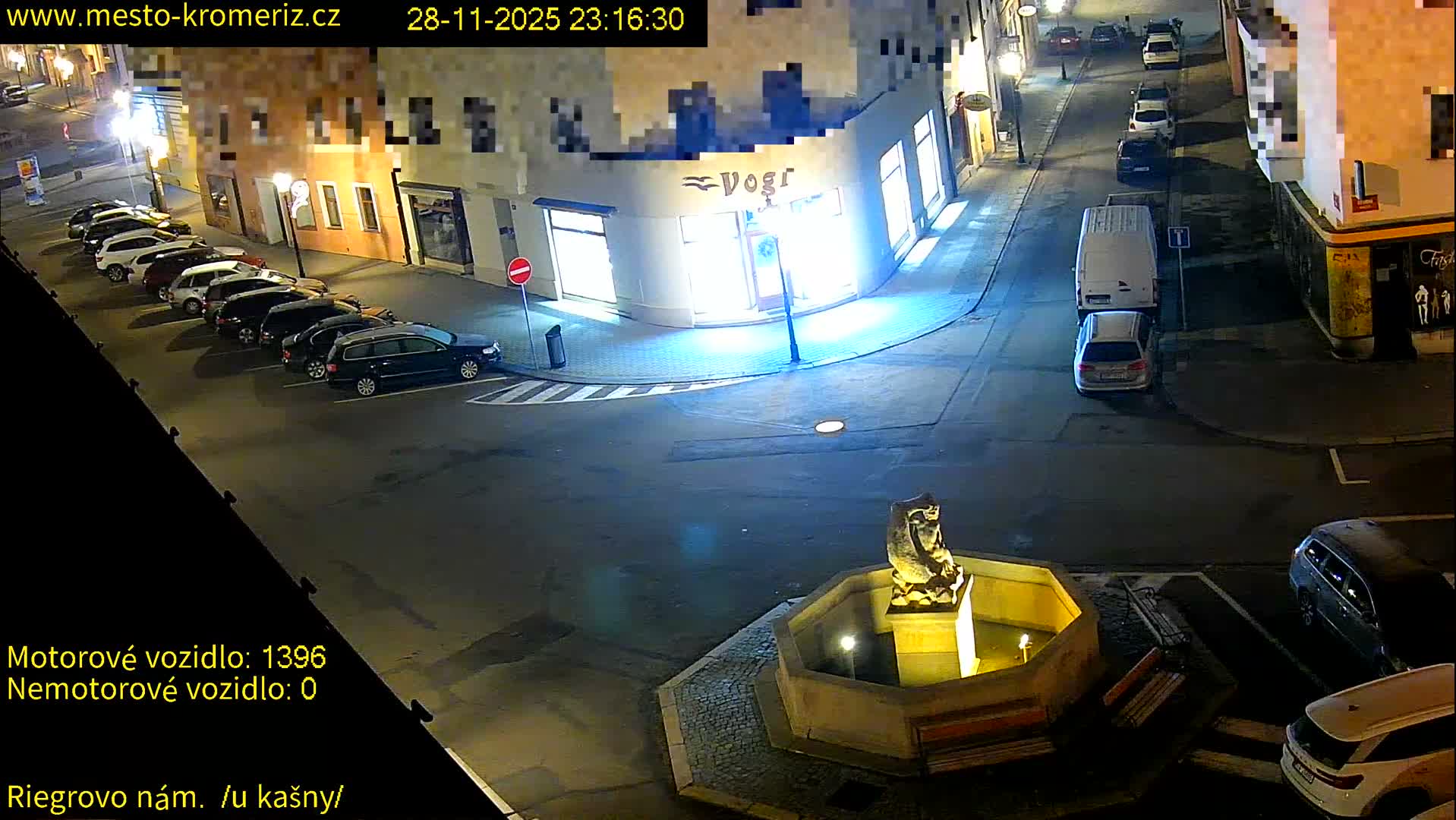 An overhead nighttime view captures a quiet urban square and intersecting streets, filled with parked cars, brightly illuminated buildings including a corner shop, and a central stone fountain, all under clear conditions.