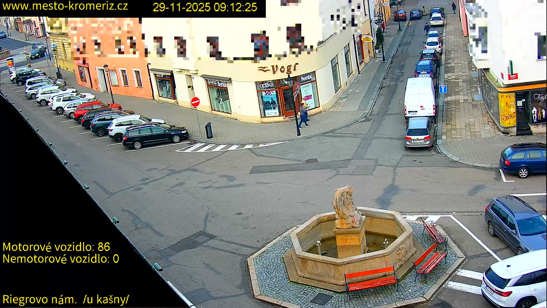 The image captures an elevated view of a bustling urban intersection on a clear day, featuring a stone fountain with benches in the foreground, numerous cars parked along the sides of paved streets, and multi-story buildings lining the roads with a few pedestrians walking on the sidewalks.