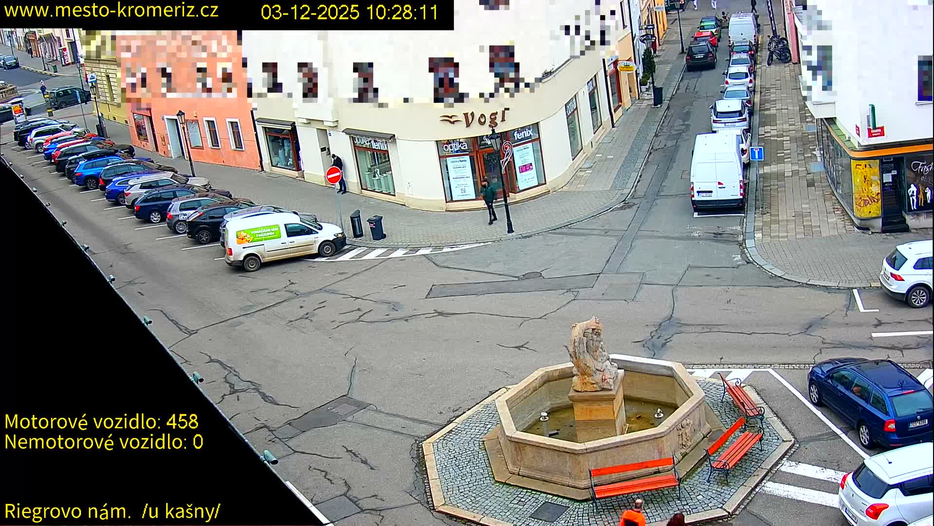 An elevated view captures a European town square on a clear day, featuring a stone fountain and benches, numerous cars parked and driving on surrounding streets, and a few pedestrians walking past shops.