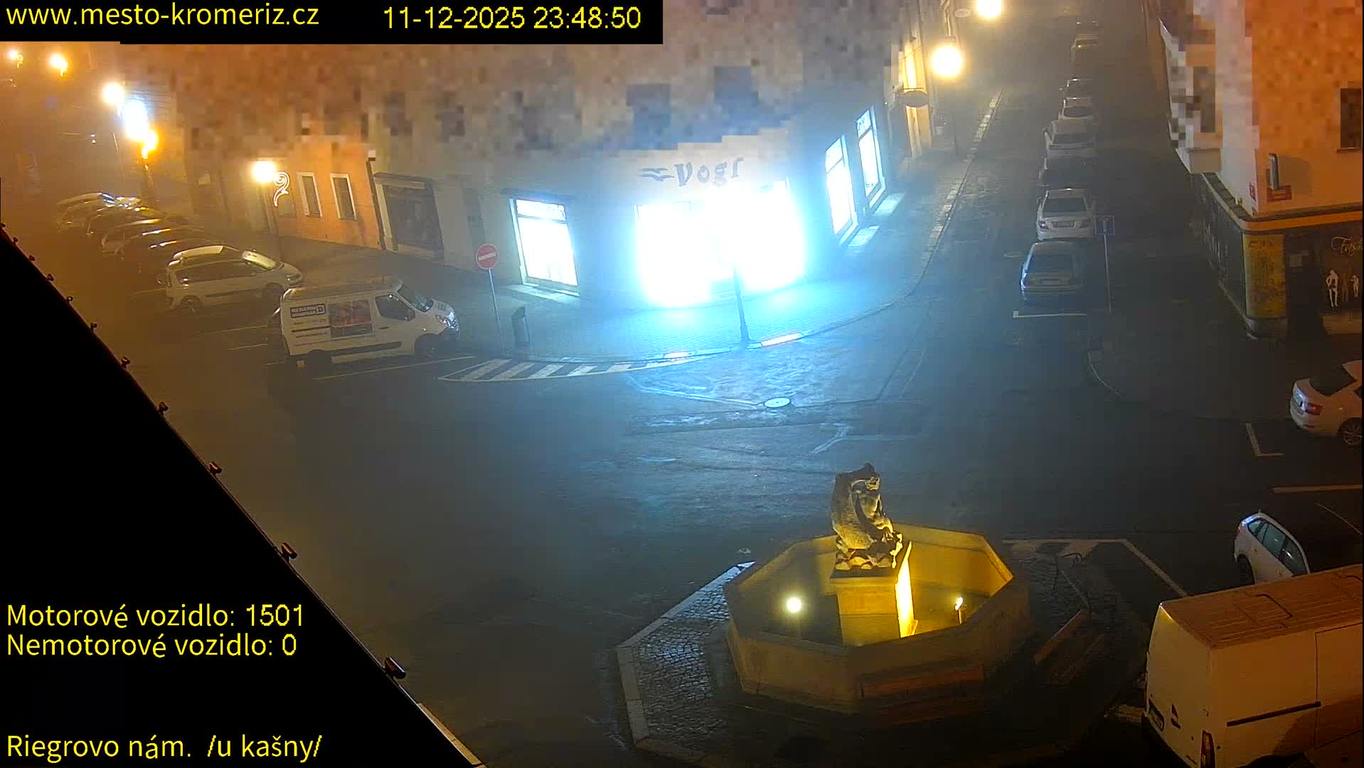 The image shows a foggy or misty urban street scene at night, illuminated by streetlights and building lights, with several parked cars and a lit fountain featuring a statue.