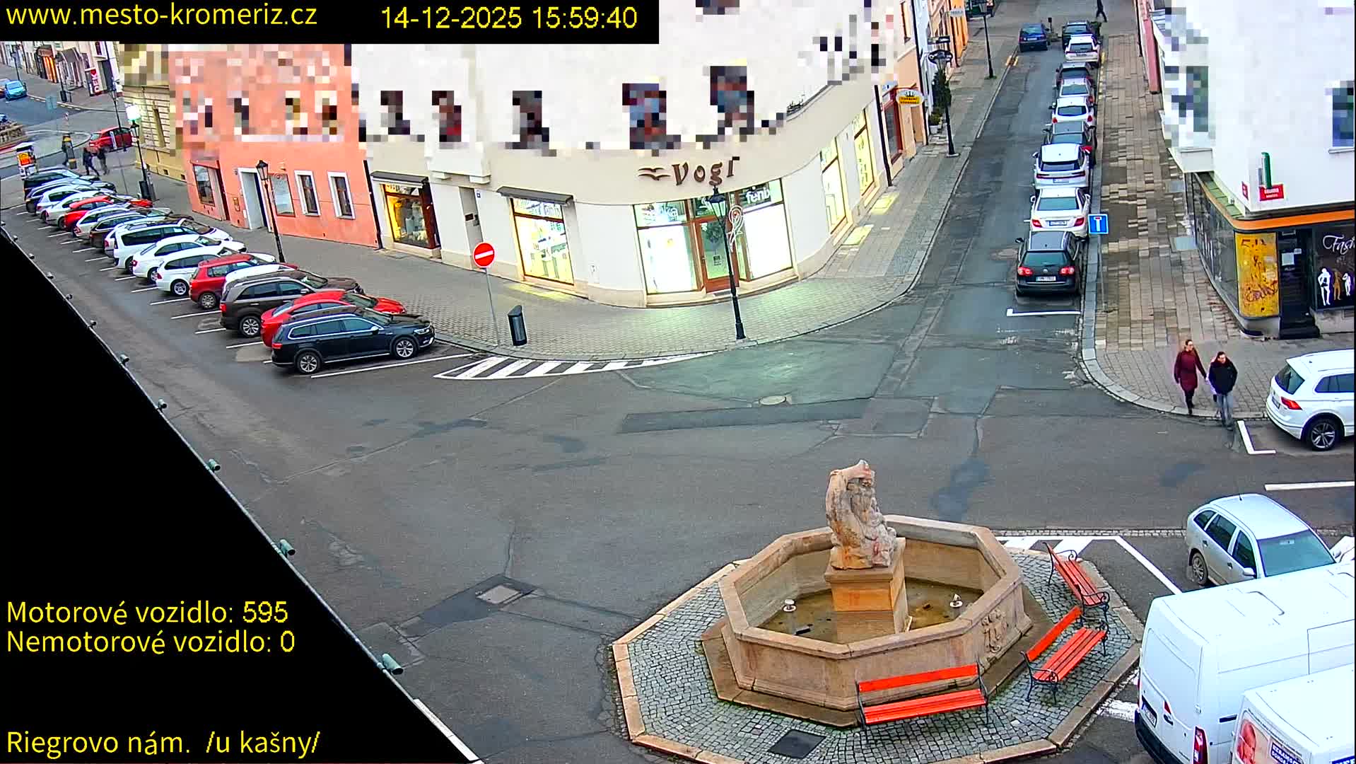 The image shows a foggy or misty urban street scene at night, illuminated by streetlights and building lights, with several parked cars and a lit fountain featuring a statue.