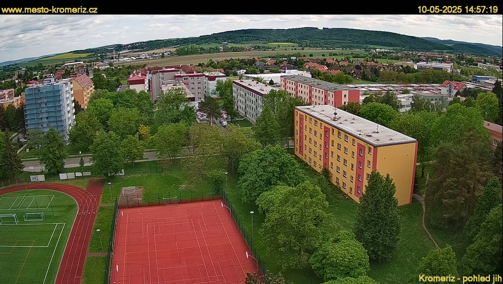 An aerial view shows several apartment buildings surrounded by lush green trees and a red sports court with a running track, under partly cloudy skies.