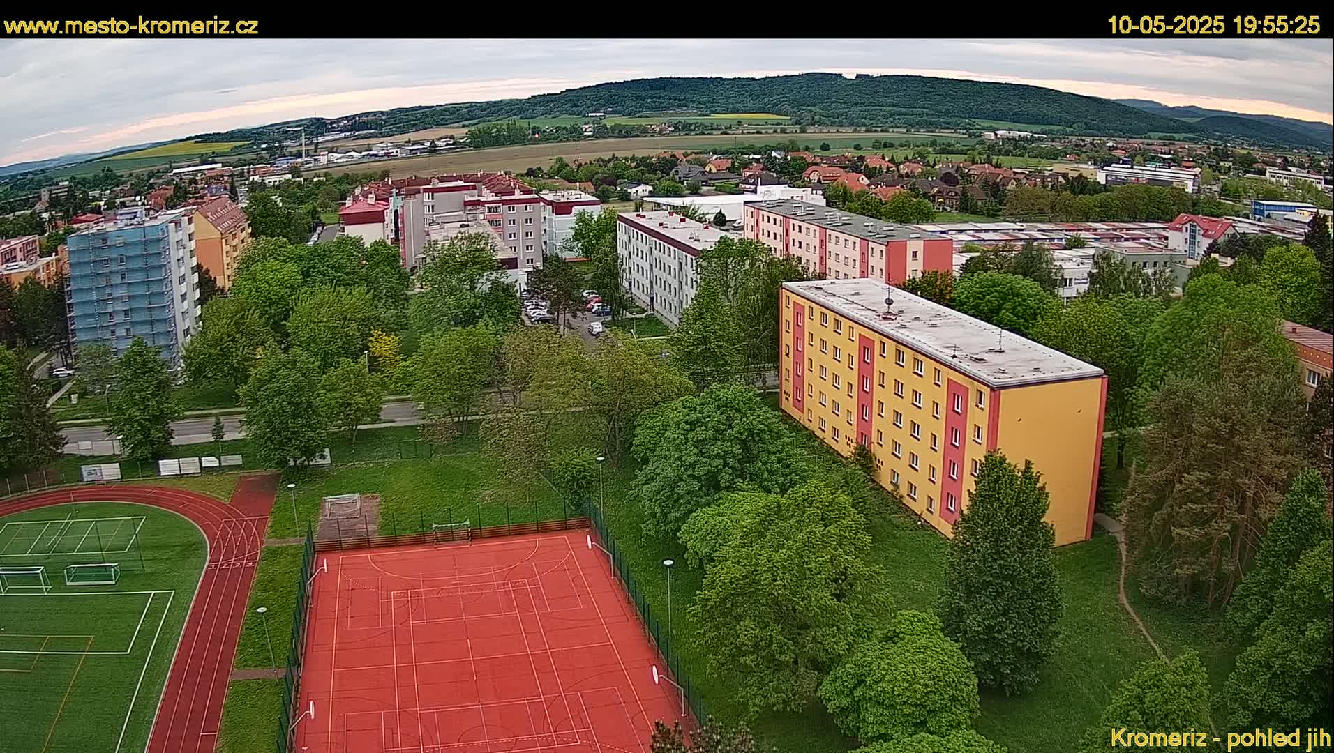 An aerial view shows multi-story apartment buildings surrounded by lush green trees and a red sports court with a running track, under an overcast sky.