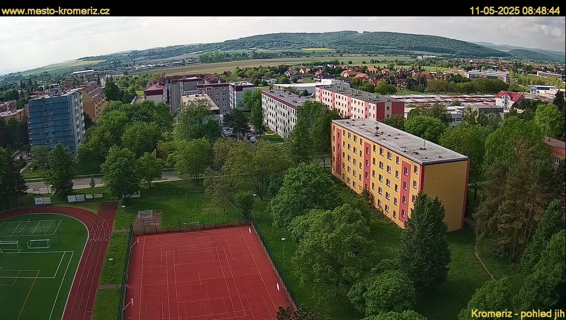 An aerial view shows apartment buildings surrounded by lush green trees and a sports field with a running track and tennis courts under a partly cloudy sky.