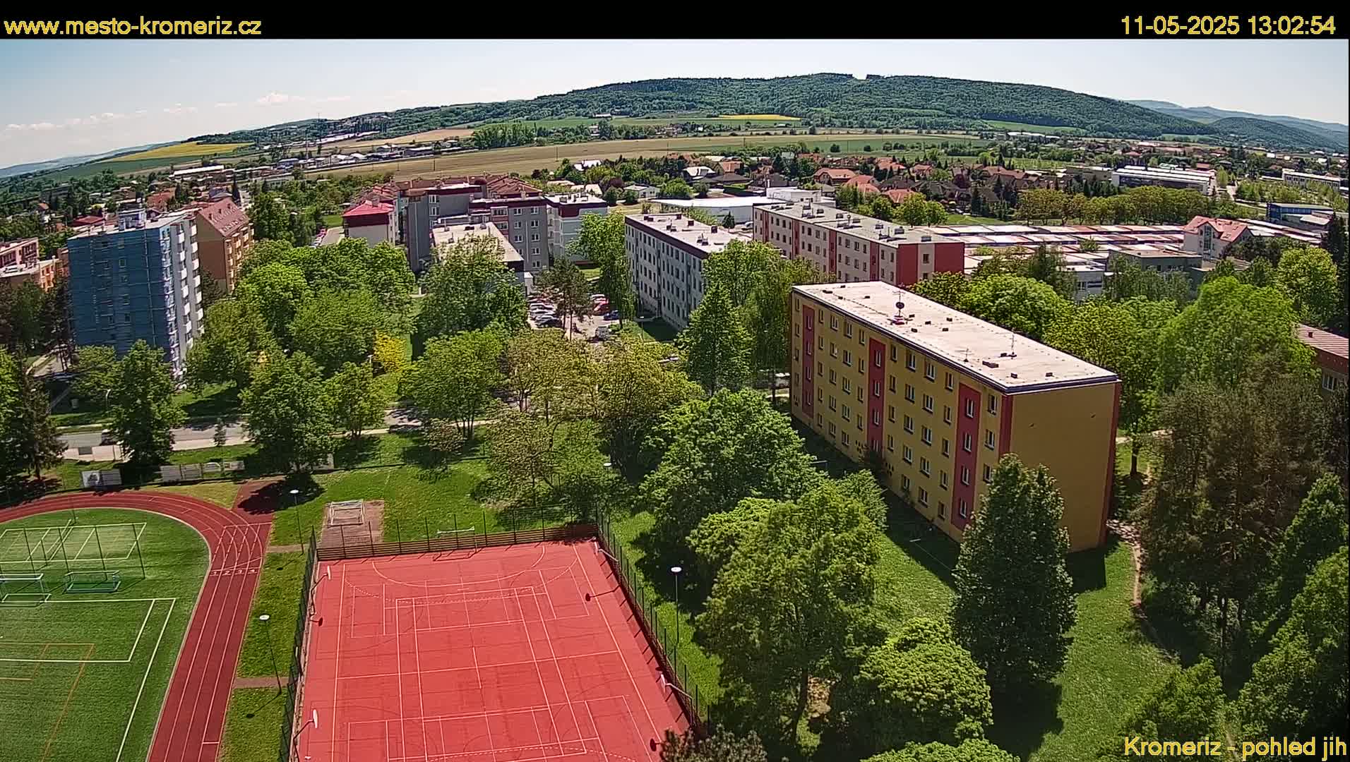An aerial view shows apartment buildings surrounded by lush green trees and a red sports court next to a running track on a sunny day.