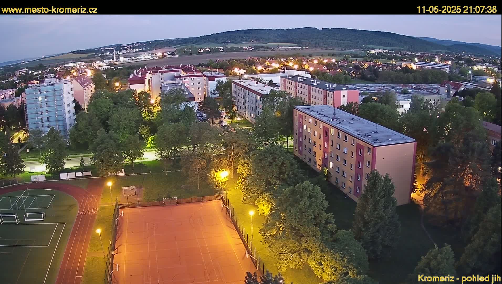 An aerial view at dusk shows a residential area with apartment buildings, a sports field with a running track, and surrounding green spaces under a clear sky.