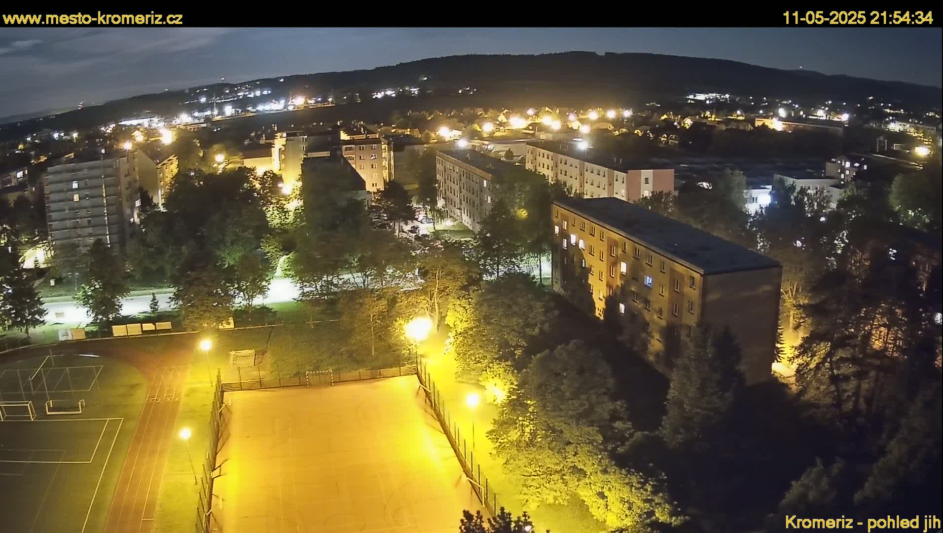 An aerial nighttime view of a residential area with a lit-up sports field, surrounded by trees and apartment buildings under a clear, dark sky.