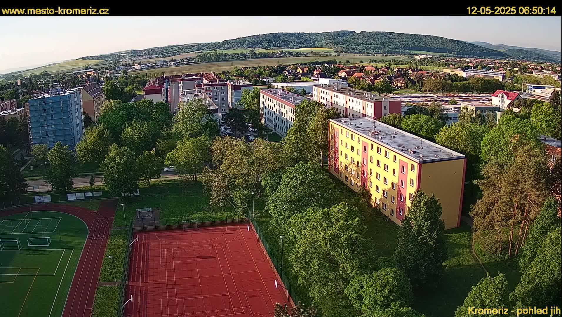 An aerial view shows apartment buildings surrounded by lush green trees and a schoolyard with athletic fields under a clear, bright sky.