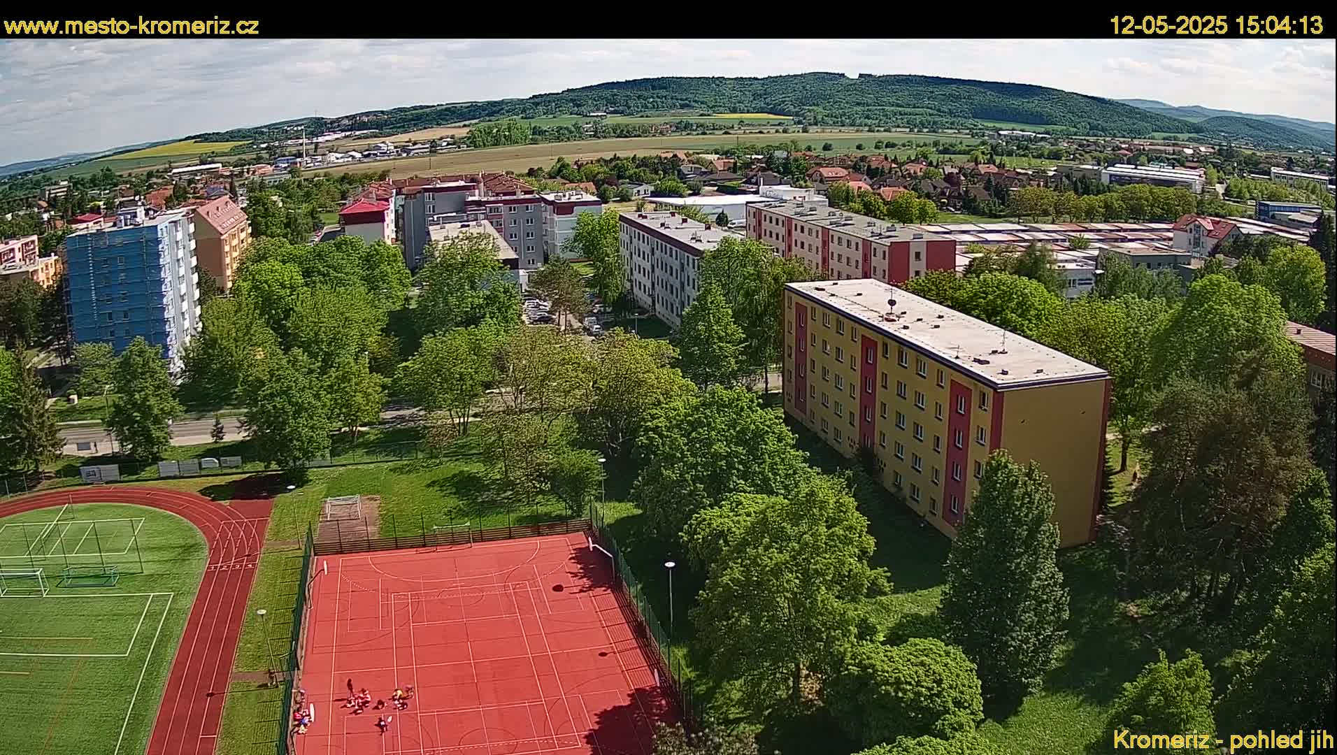 An aerial view shows a residential area with apartment buildings surrounded by lush green trees, a red sports court, and a running track under a partly sunny sky.