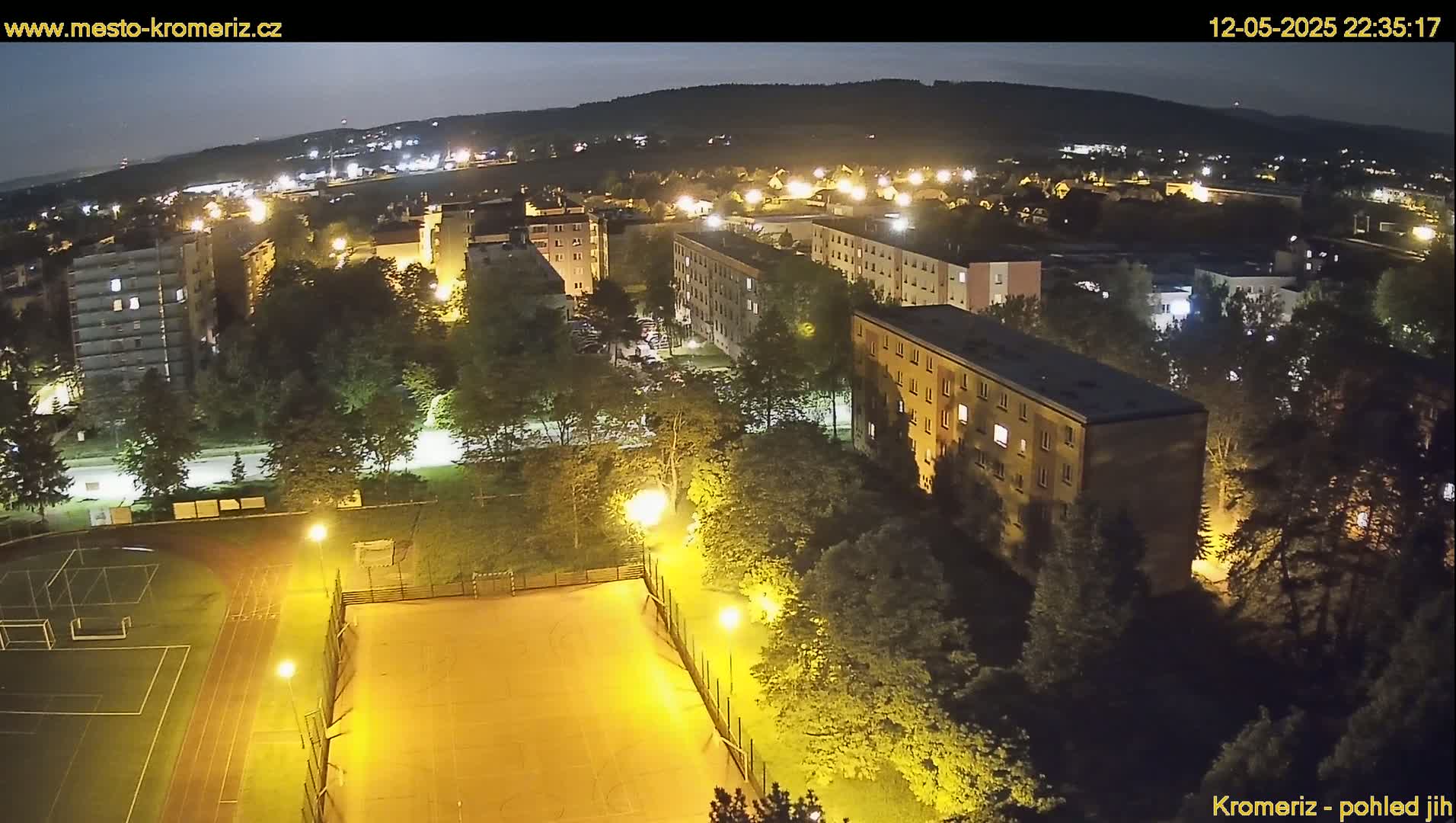 An aerial nighttime view of a residential area with a sports field illuminated by bright lights, under a dark sky.