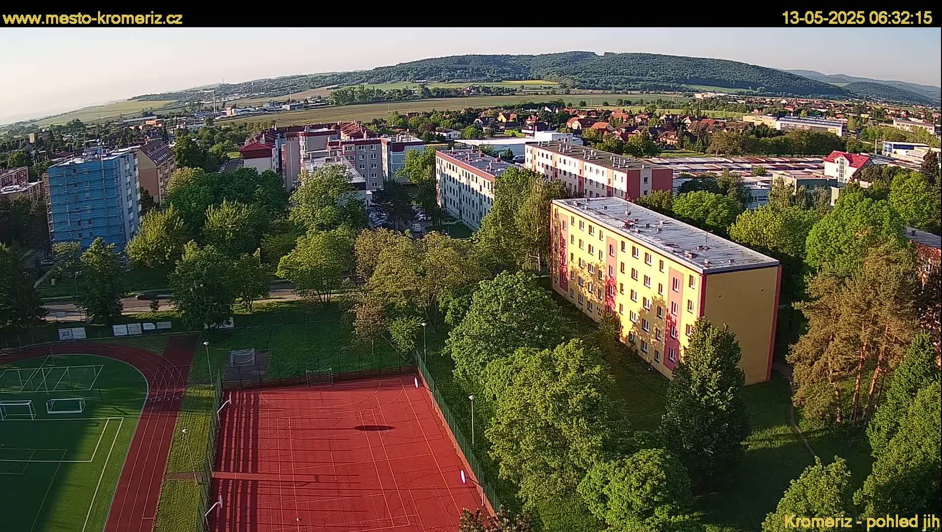 An aerial view shows several apartment buildings surrounded by lush green trees and a red sports court under a clear sky.