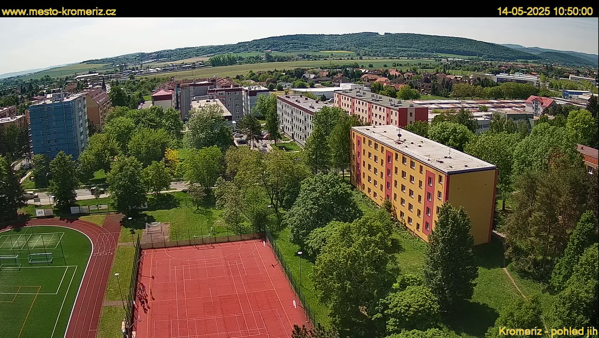 An aerial view shows apartment buildings, green trees, a red sports court, and a running track on a sunny day.