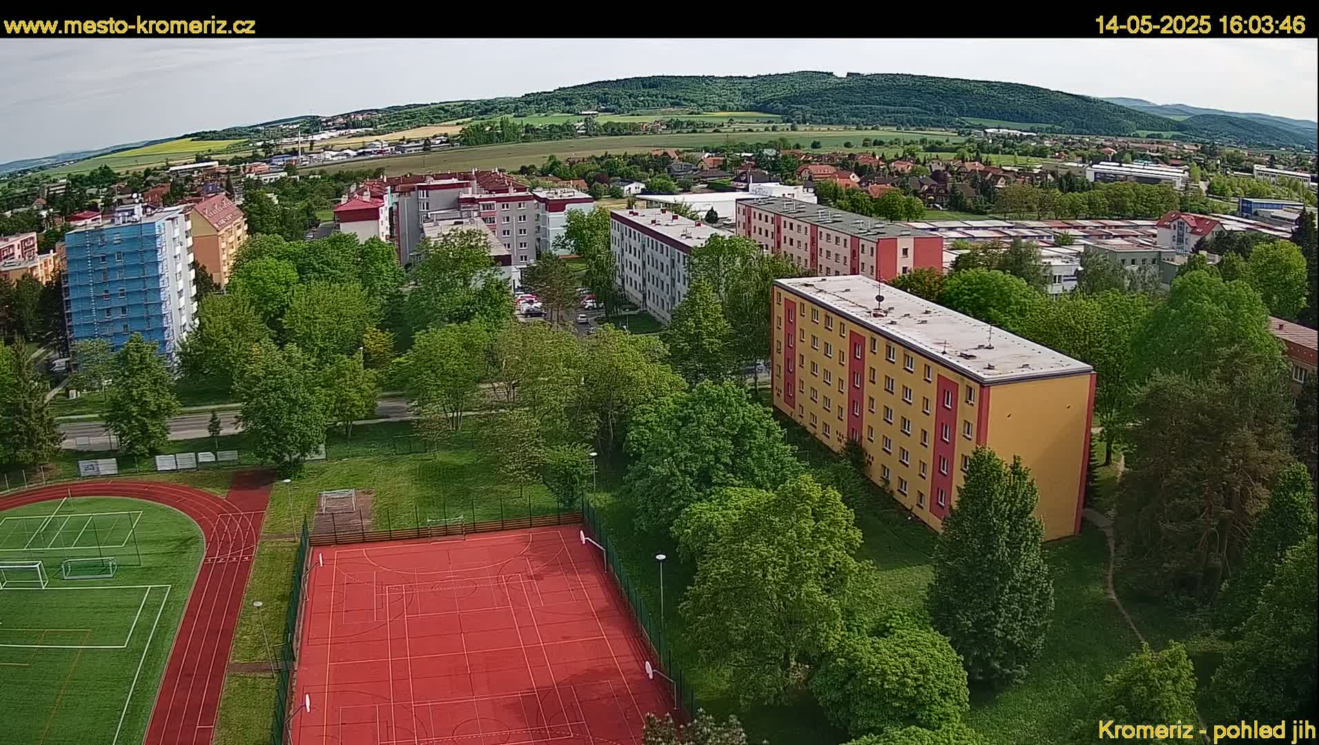 An aerial view shows a residential area with apartment buildings, green trees, a running track, and a multi-purpose sports court on a partly sunny day.