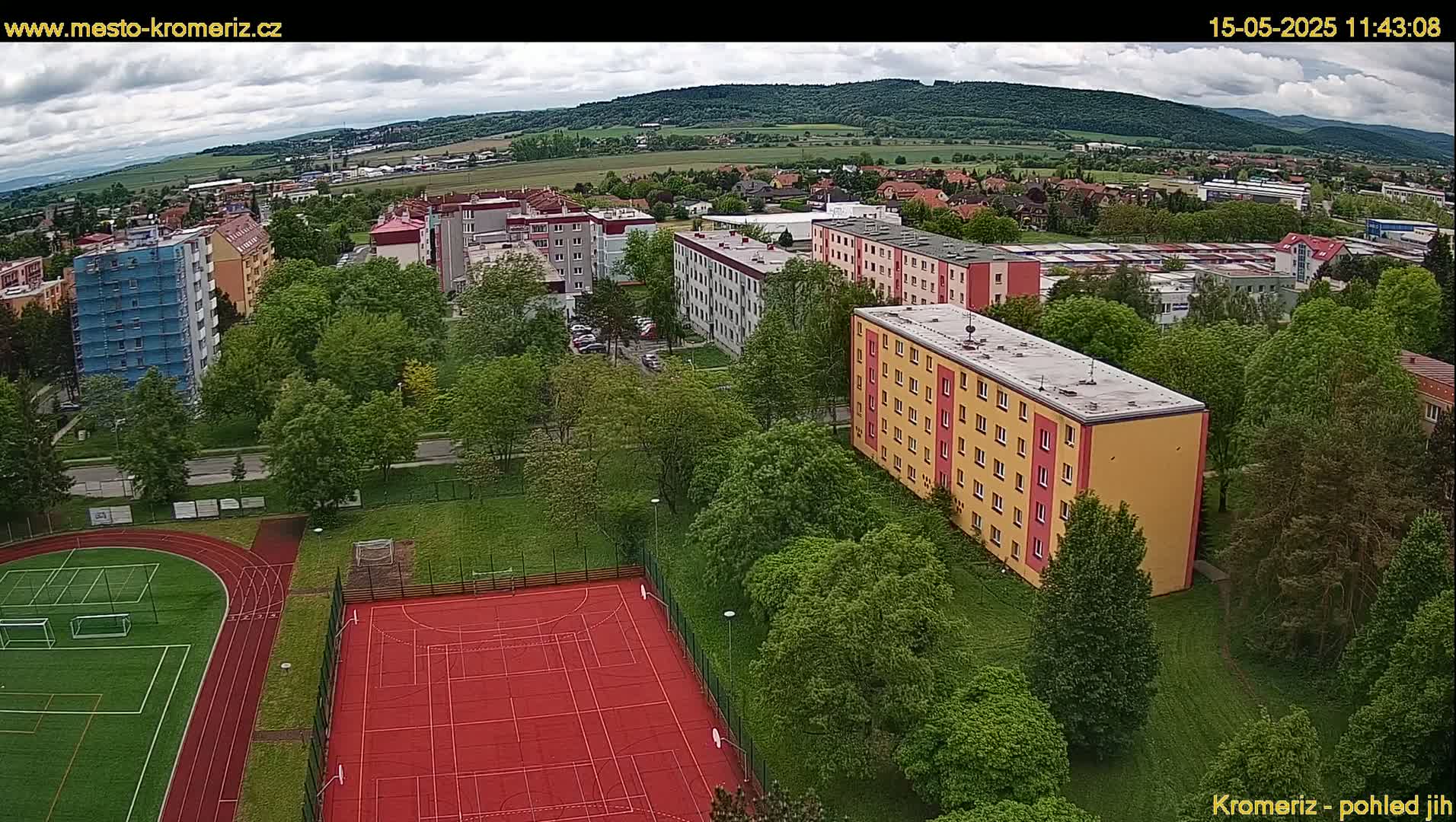 An aerial view shows a residential area with apartment buildings, green trees, and a red sports court with a running track, under partly cloudy skies.