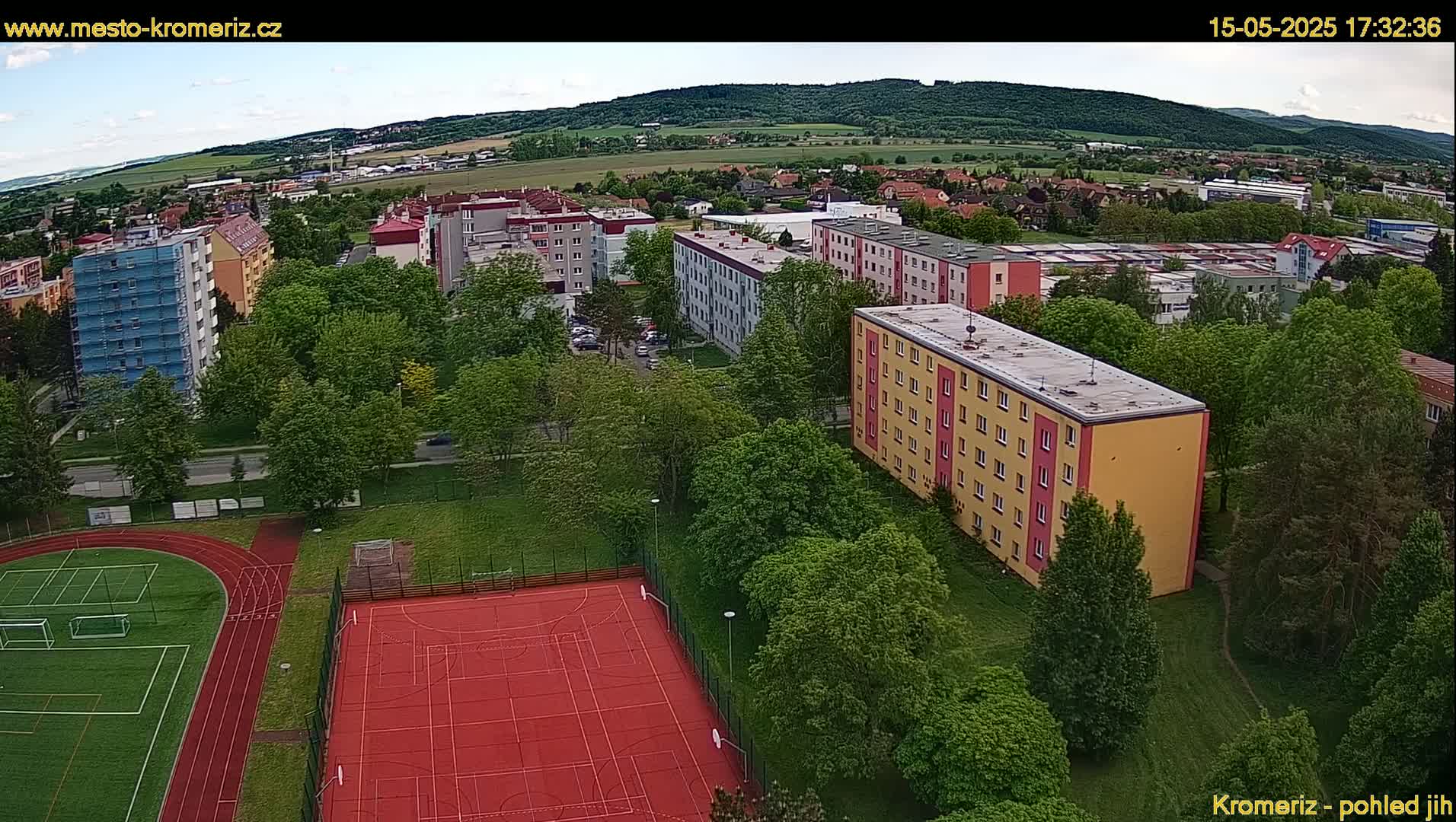 An aerial view shows a residential area with several apartment buildings, a running track, and a multi-purpose sports court, all surrounded by lush green trees under a partly cloudy sky.