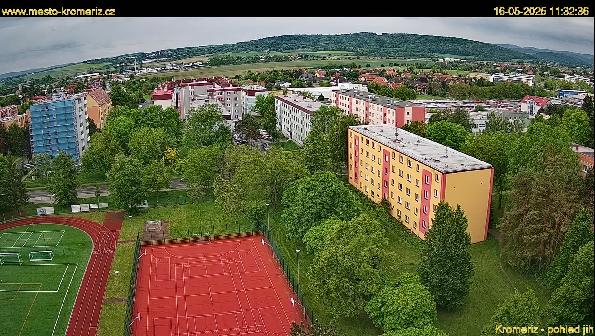 An aerial view shows an overcast day over a residential area with green trees and a red athletic field, including a running track and a soccer field.