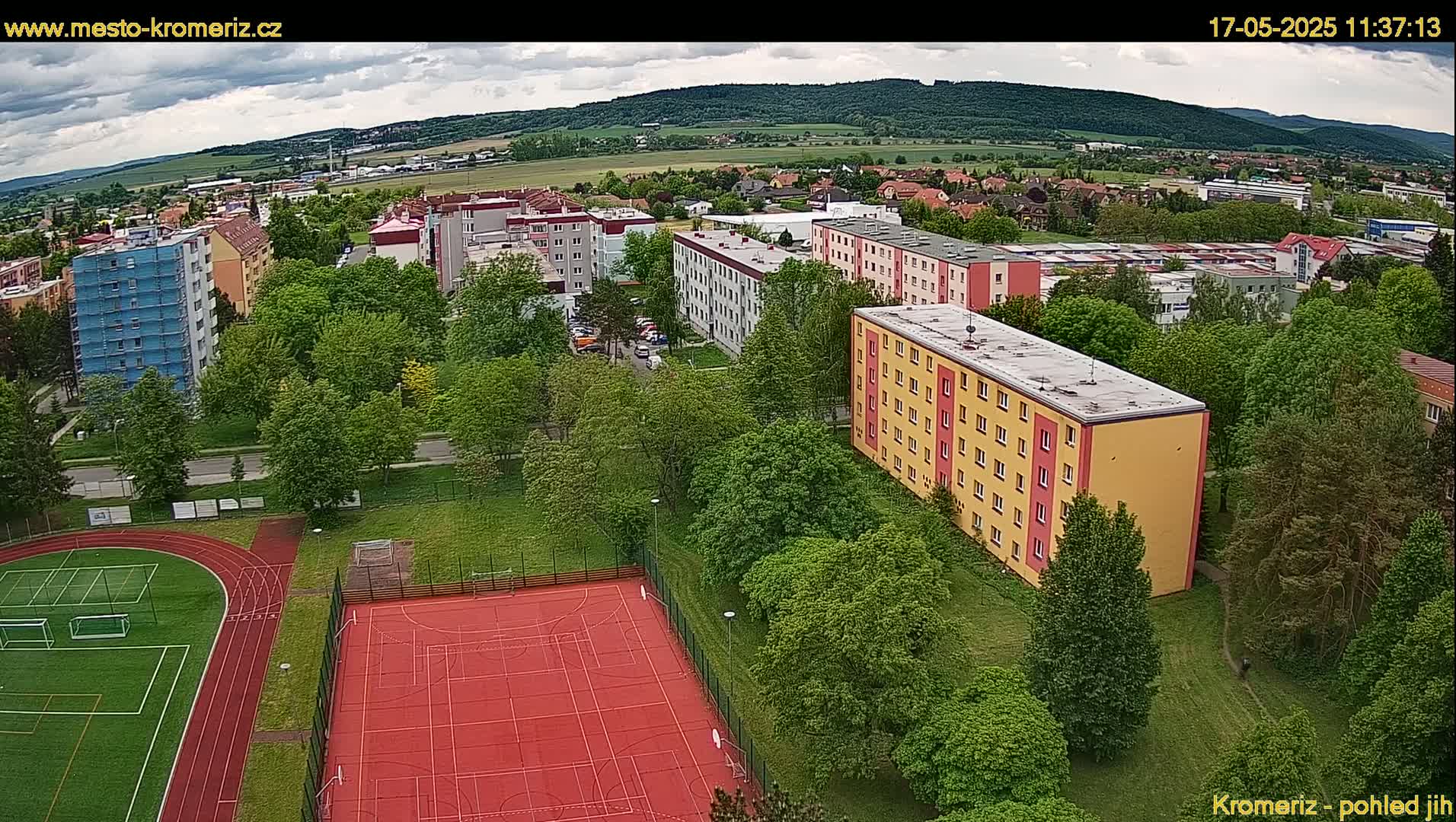 An aerial view shows apartment buildings surrounded by lush green trees and a sports field with a running track and basketball court under partly cloudy skies.