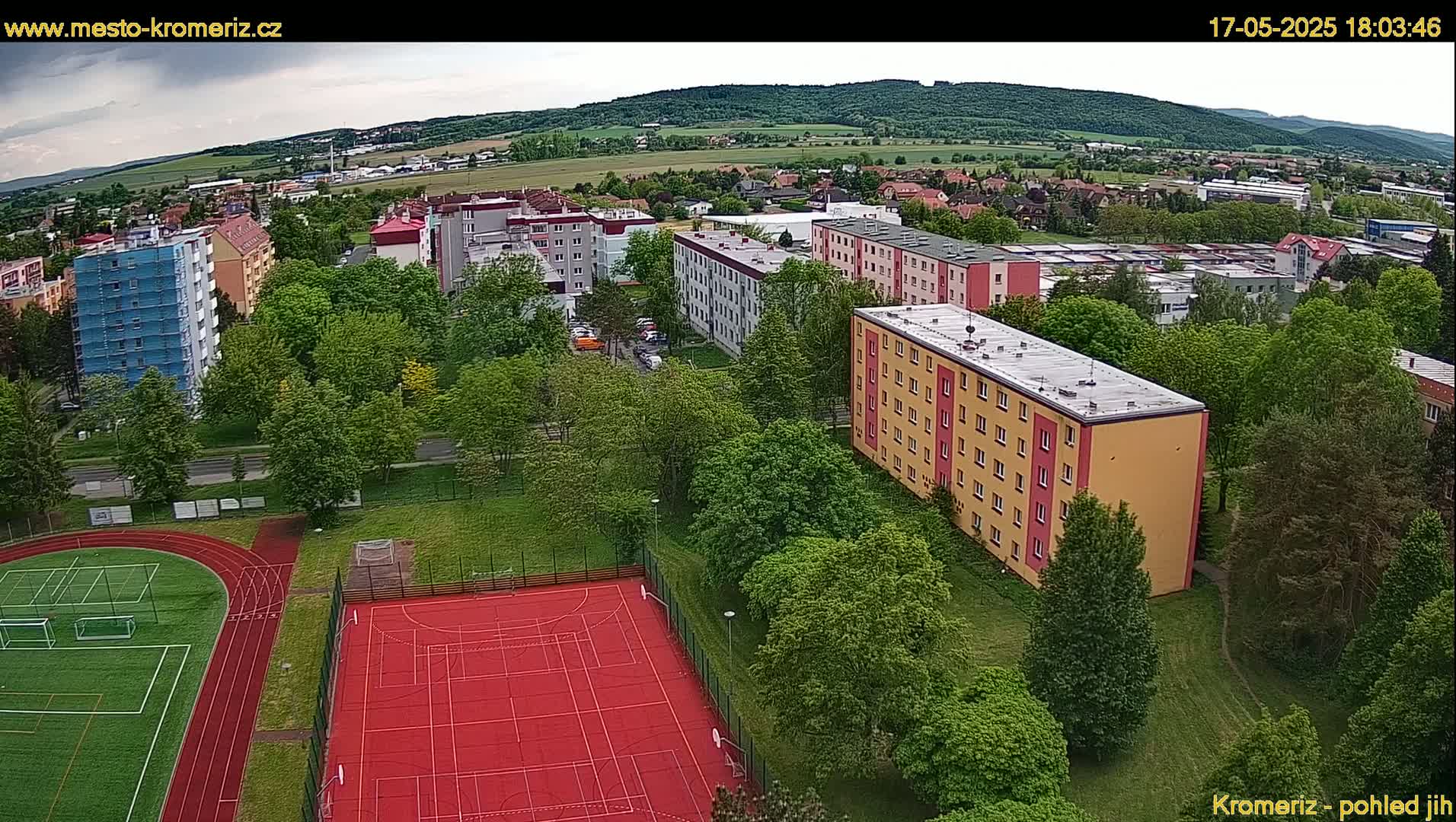 An aerial view shows apartment buildings, green trees, and a red outdoor sports court under a partly cloudy sky.