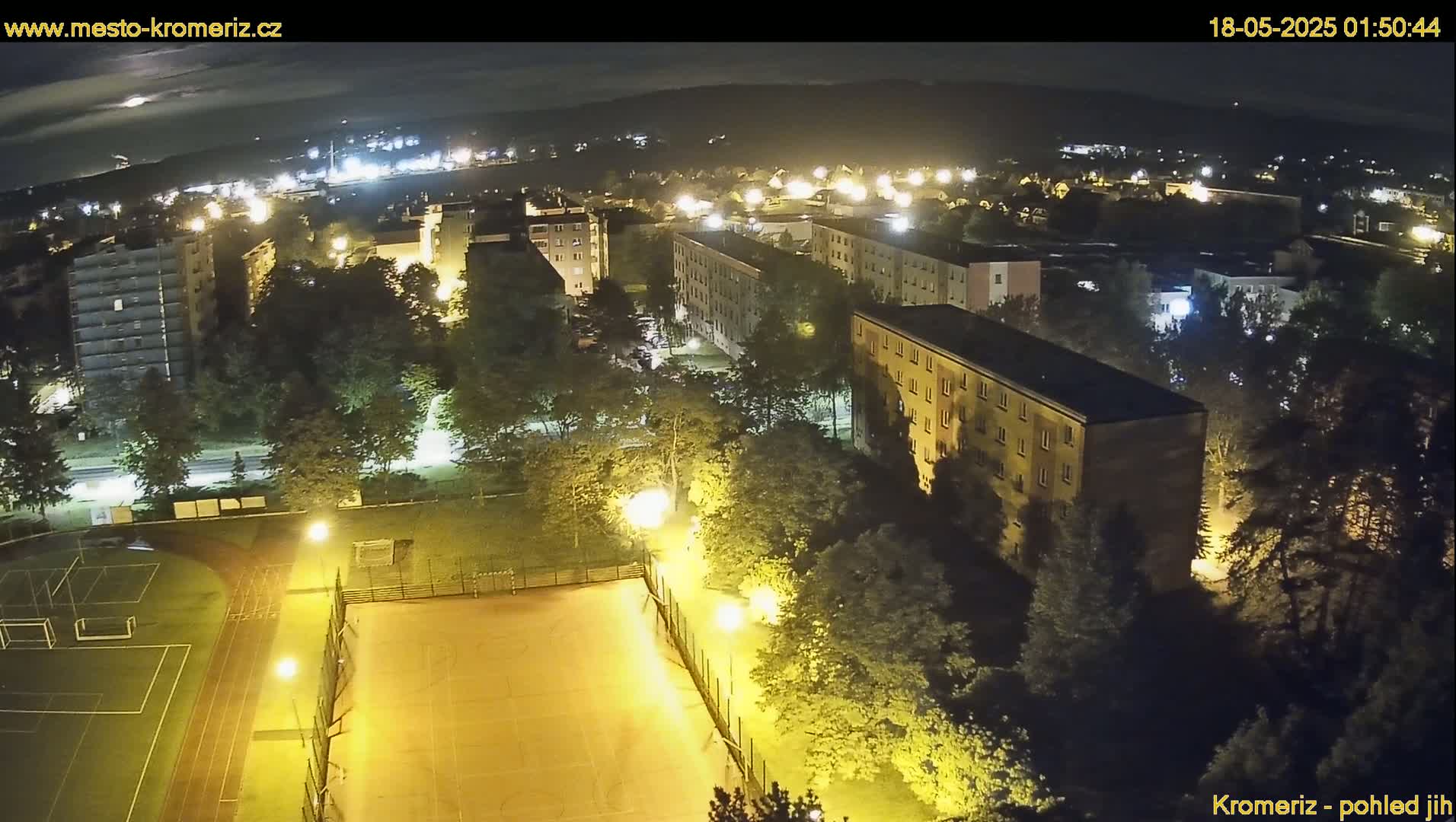 A nighttime aerial view shows a residential area with multi-story buildings, a sports field illuminated by artificial lights, and surrounding trees under a clear sky.