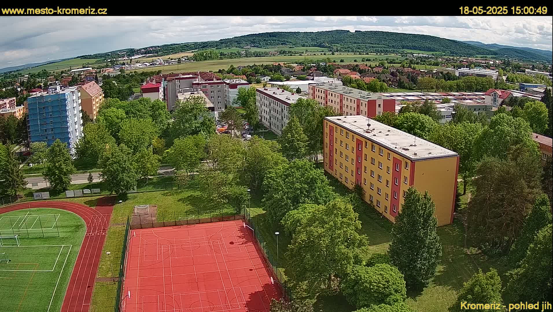 An aerial view shows a residential area with apartment buildings surrounded by lush green trees and a red-surfaced sports court with a running track under a partly cloudy sky.