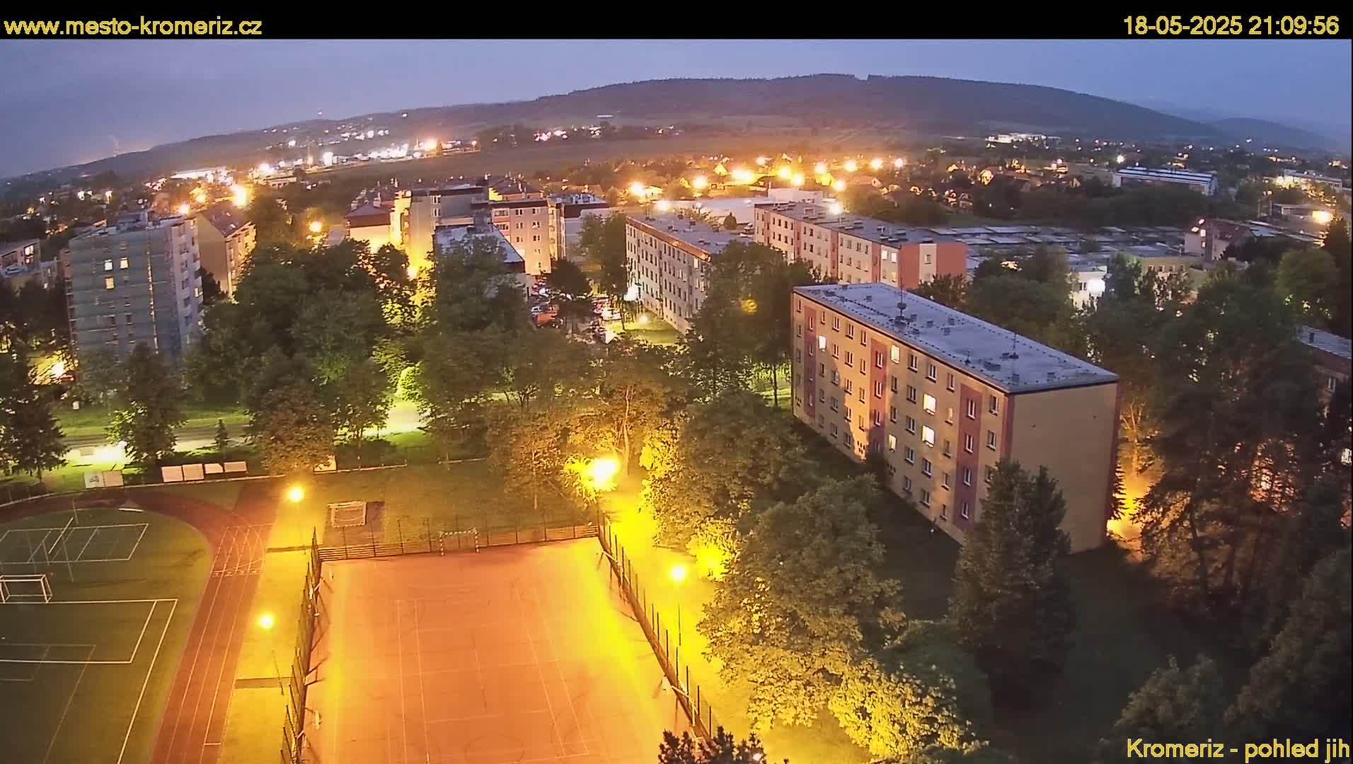 An aerial nighttime view of a residential area with illuminated apartment buildings, green spaces, and sports fields under a clear, dark sky.
