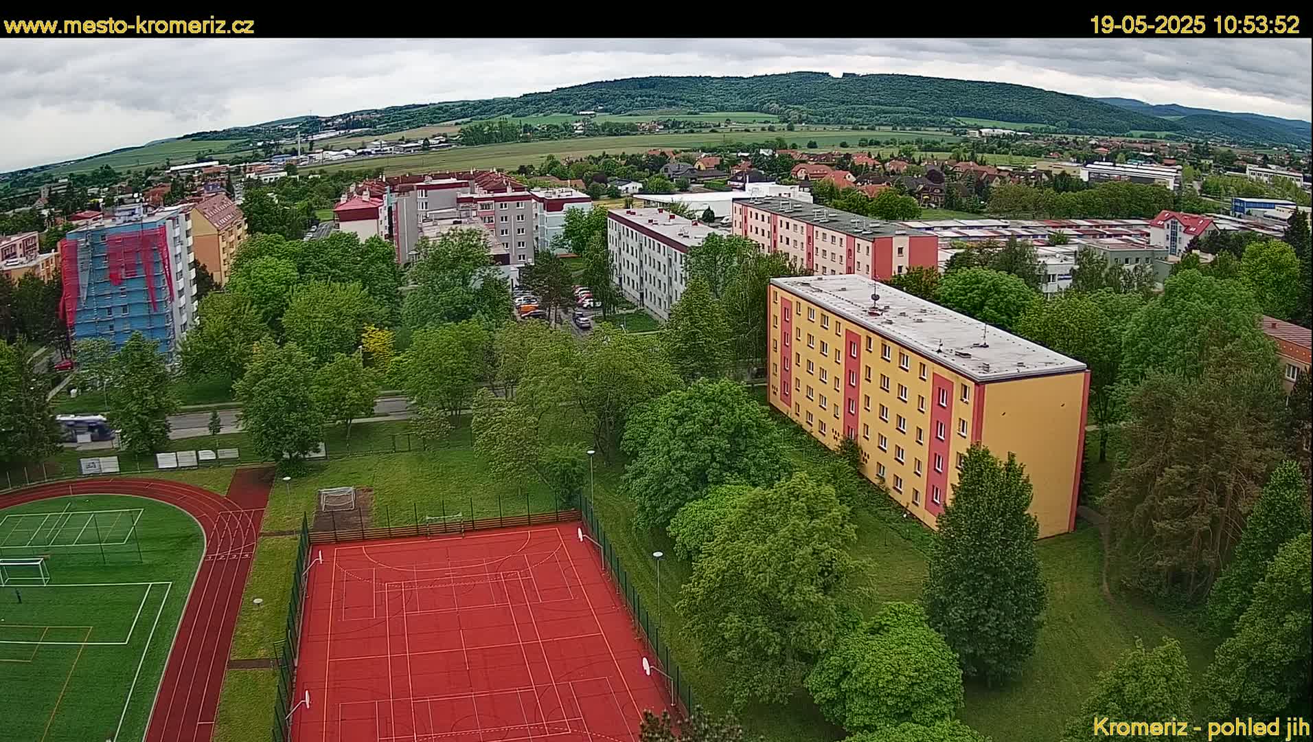 An aerial view shows a residential area with several apartment buildings surrounded by lush green trees and a red sports court, under a partly cloudy sky.
