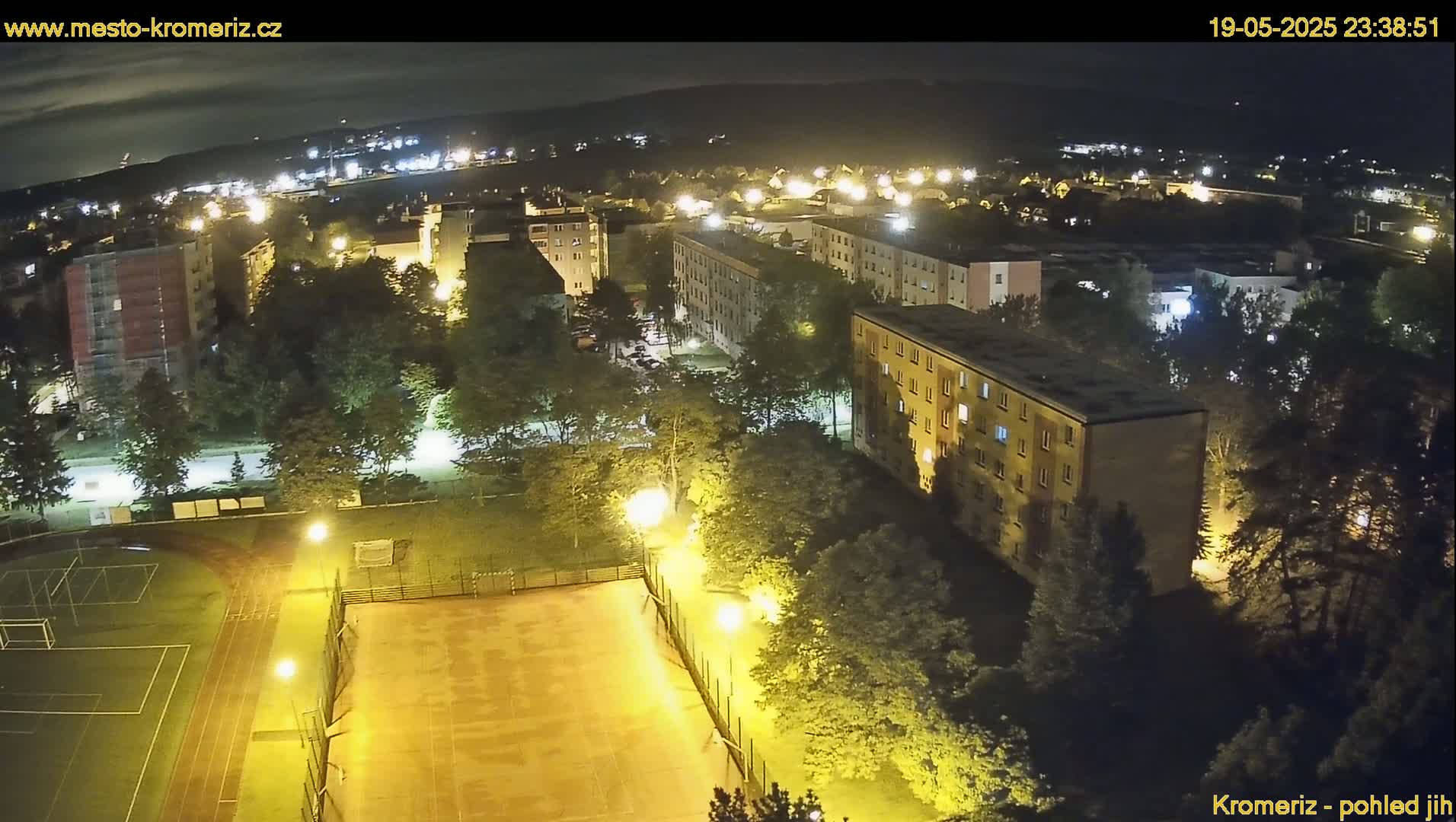 An aerial nighttime view of a residential area with a lit-up sports court and surrounding trees, under a clear sky.