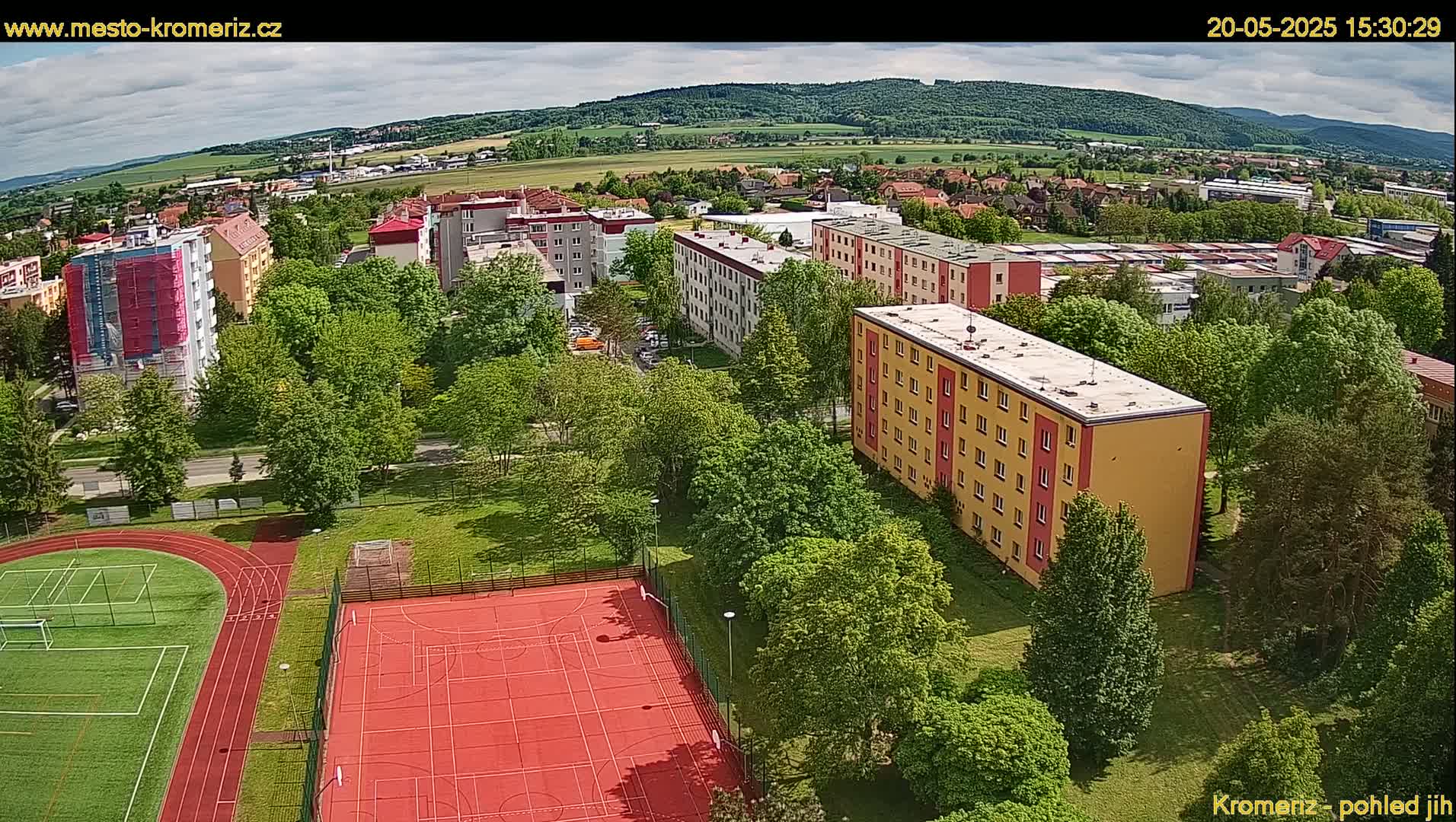 An aerial view shows apartment buildings surrounded by lush green trees and a sports field with a running track under partly cloudy skies.
