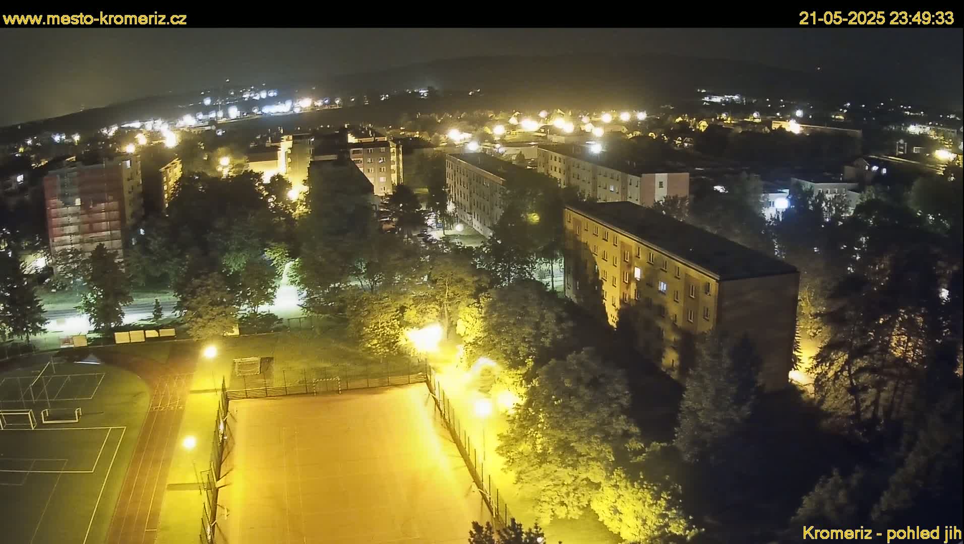 An aerial nighttime view of a residential area with illuminated apartment buildings, trees, and a sports field.