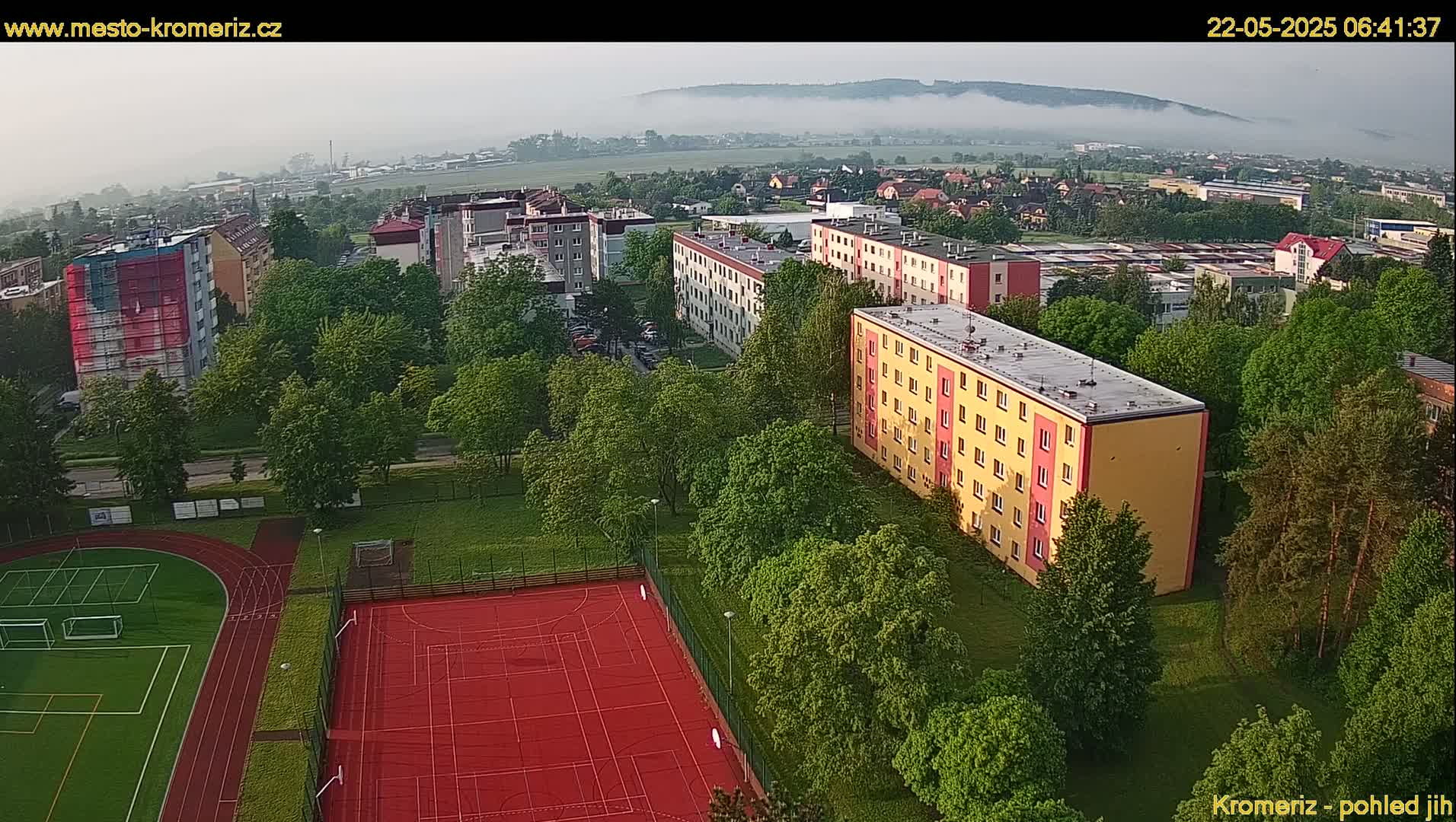 A misty morning view overlooking apartment buildings, green spaces, athletic fields, and a partially-covered building under construction.