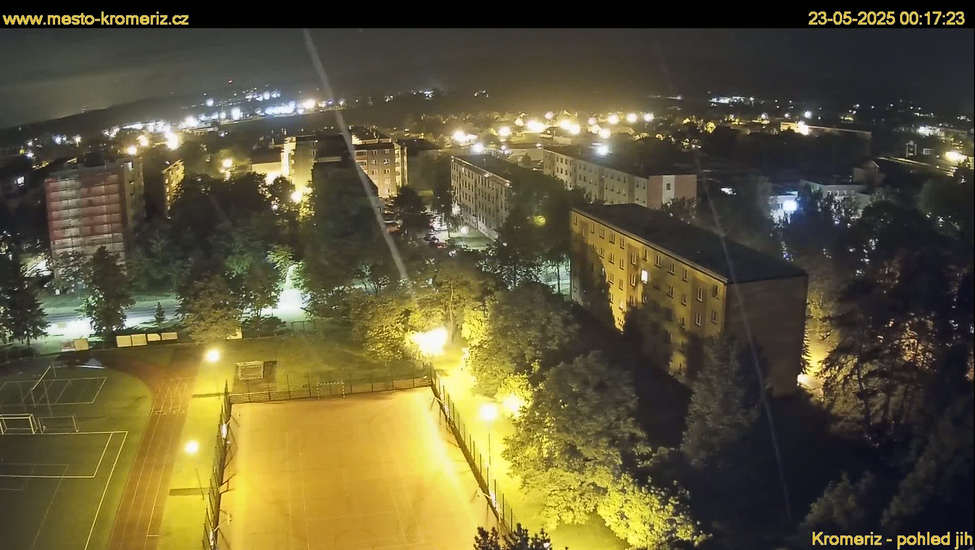 An aerial nighttime view shows a residential area with illuminated buildings, trees, and a sports field, under a clear sky.