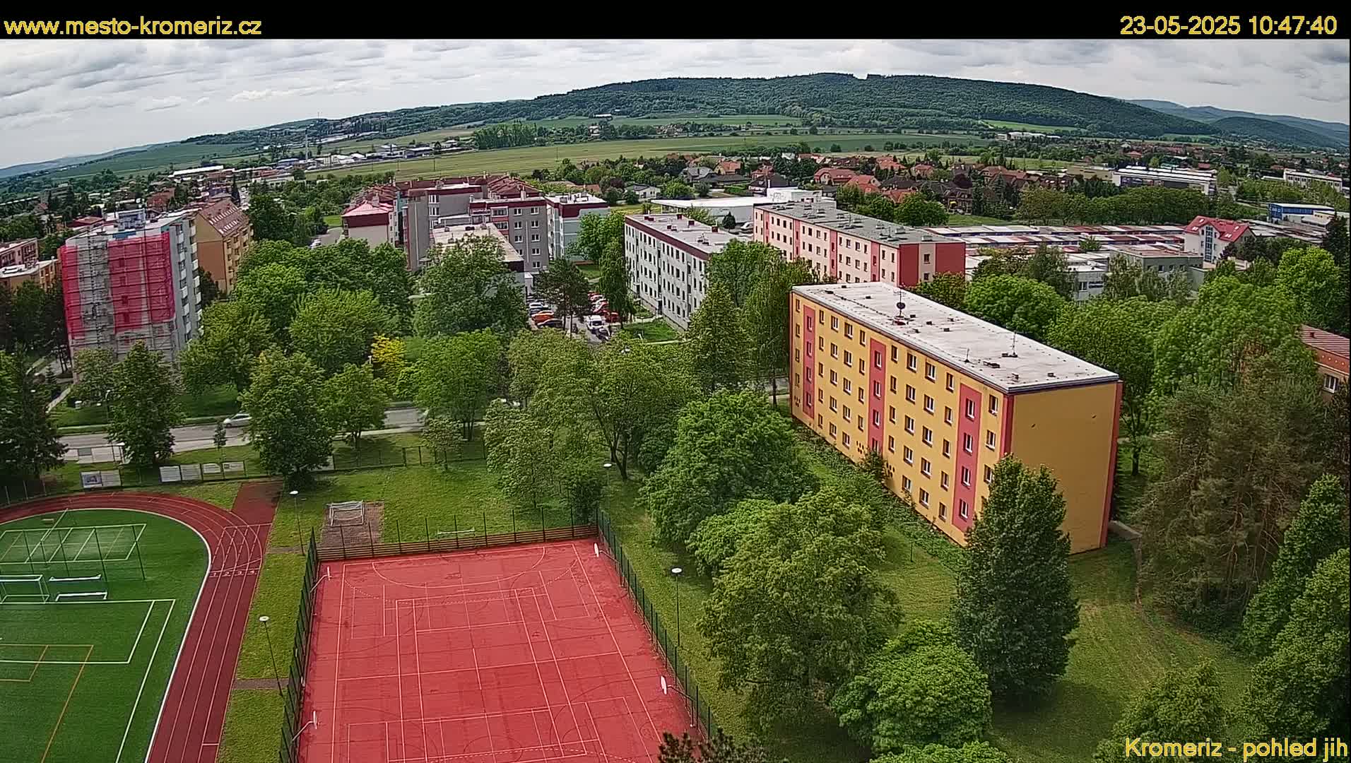 An aerial view shows multi-story apartment buildings surrounded by lush green trees and a red sports court, all under a partly cloudy sky.