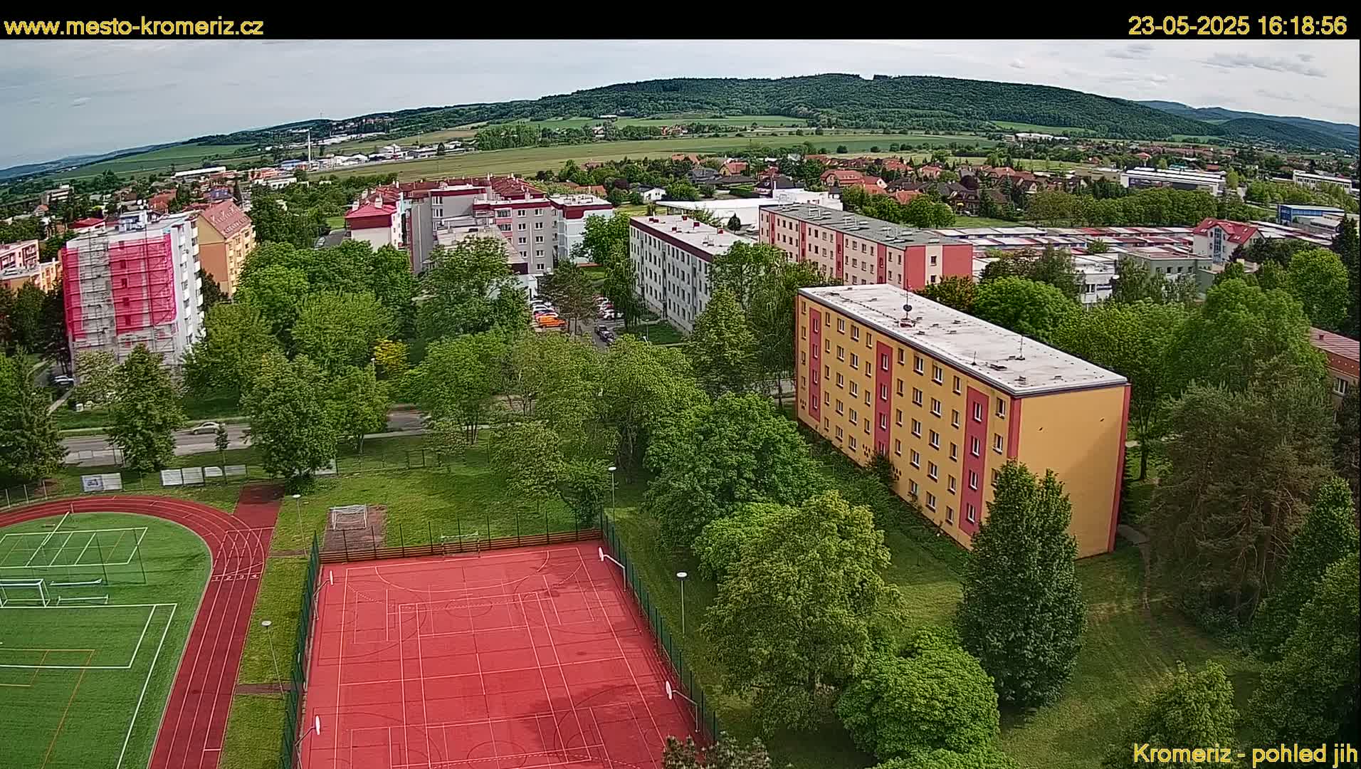 An aerial view shows a residential area with multi-story buildings, green trees, a red sports court, and a running track under a partly cloudy sky.