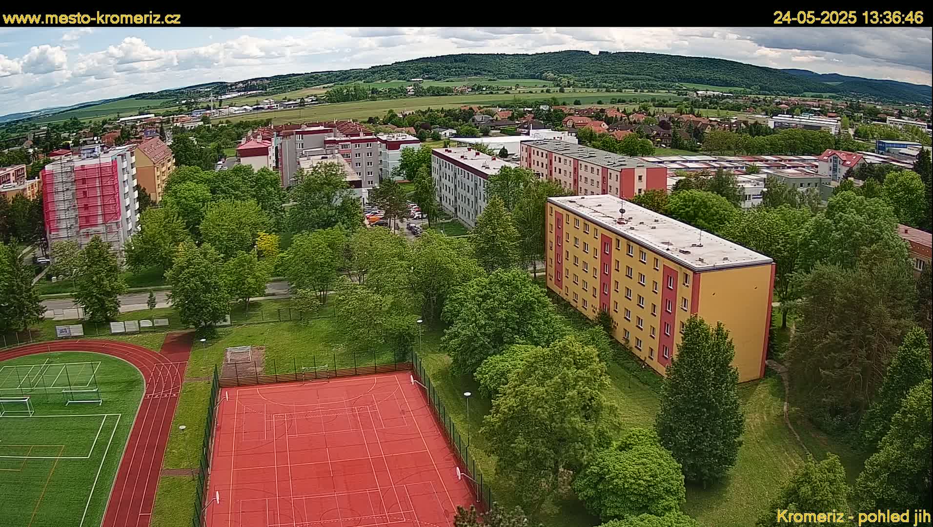 An aerial view shows a residential area with apartment buildings surrounded by lush green trees, a red outdoor basketball court, and a running track under a partly cloudy sky.