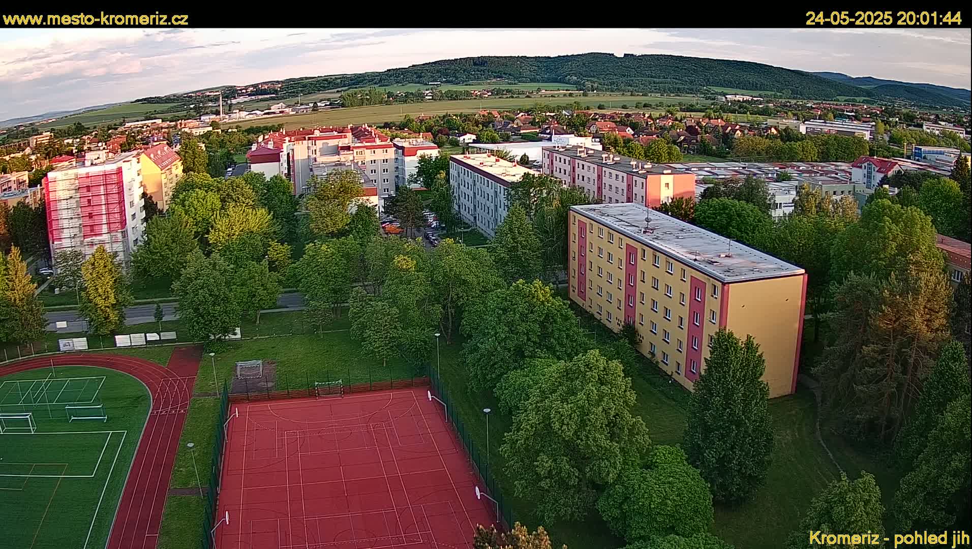 An aerial view shows apartment buildings and a sports field surrounded by lush green trees under a partly cloudy evening sky.