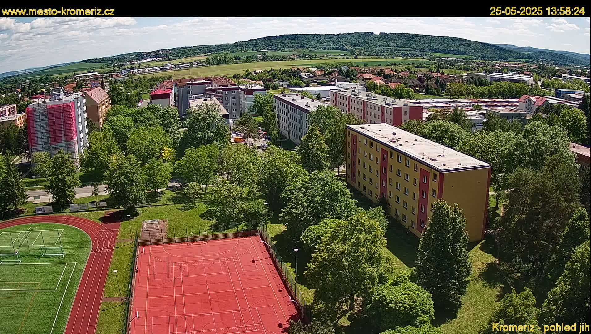 An aerial view shows a residential area with apartment buildings, green spaces, a running track, and a multi-purpose sports court under sunny skies.