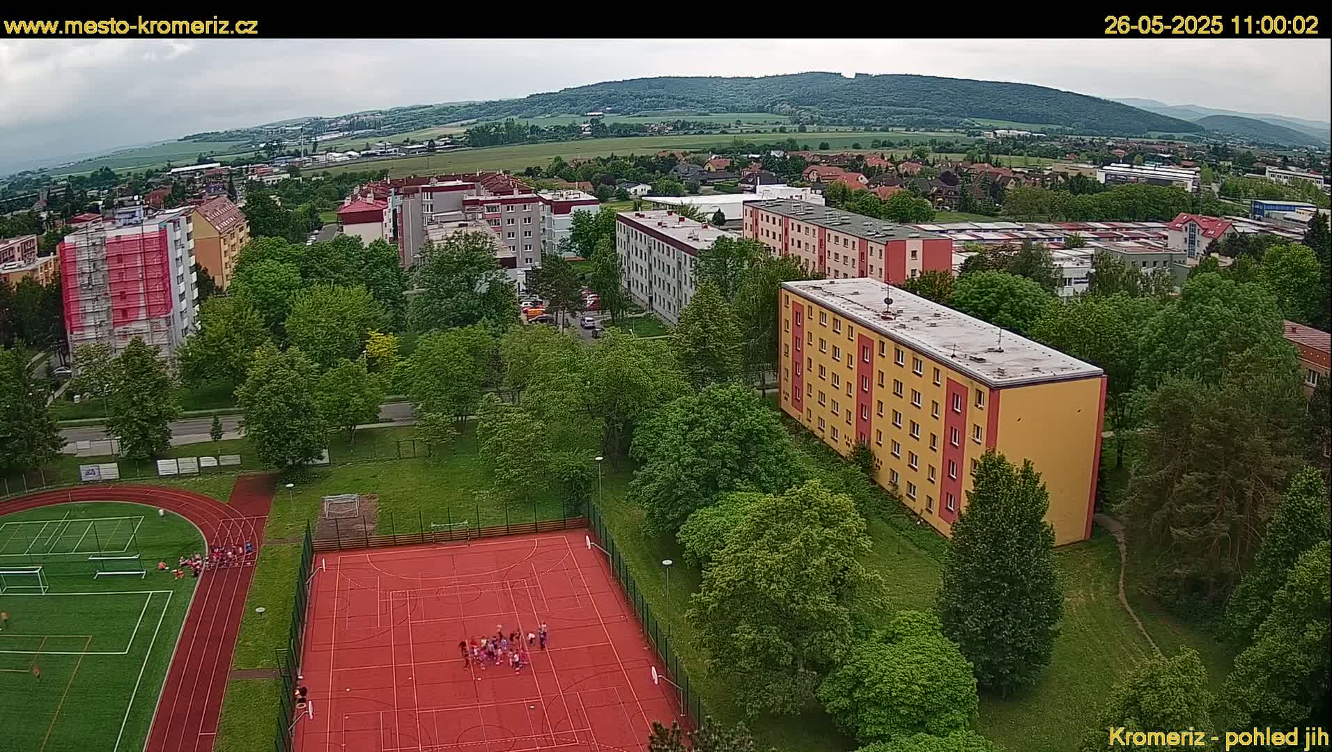 An aerial view shows a residential area with apartment buildings, lush green trees, a red sports court with people on it, and a running track under an overcast sky.